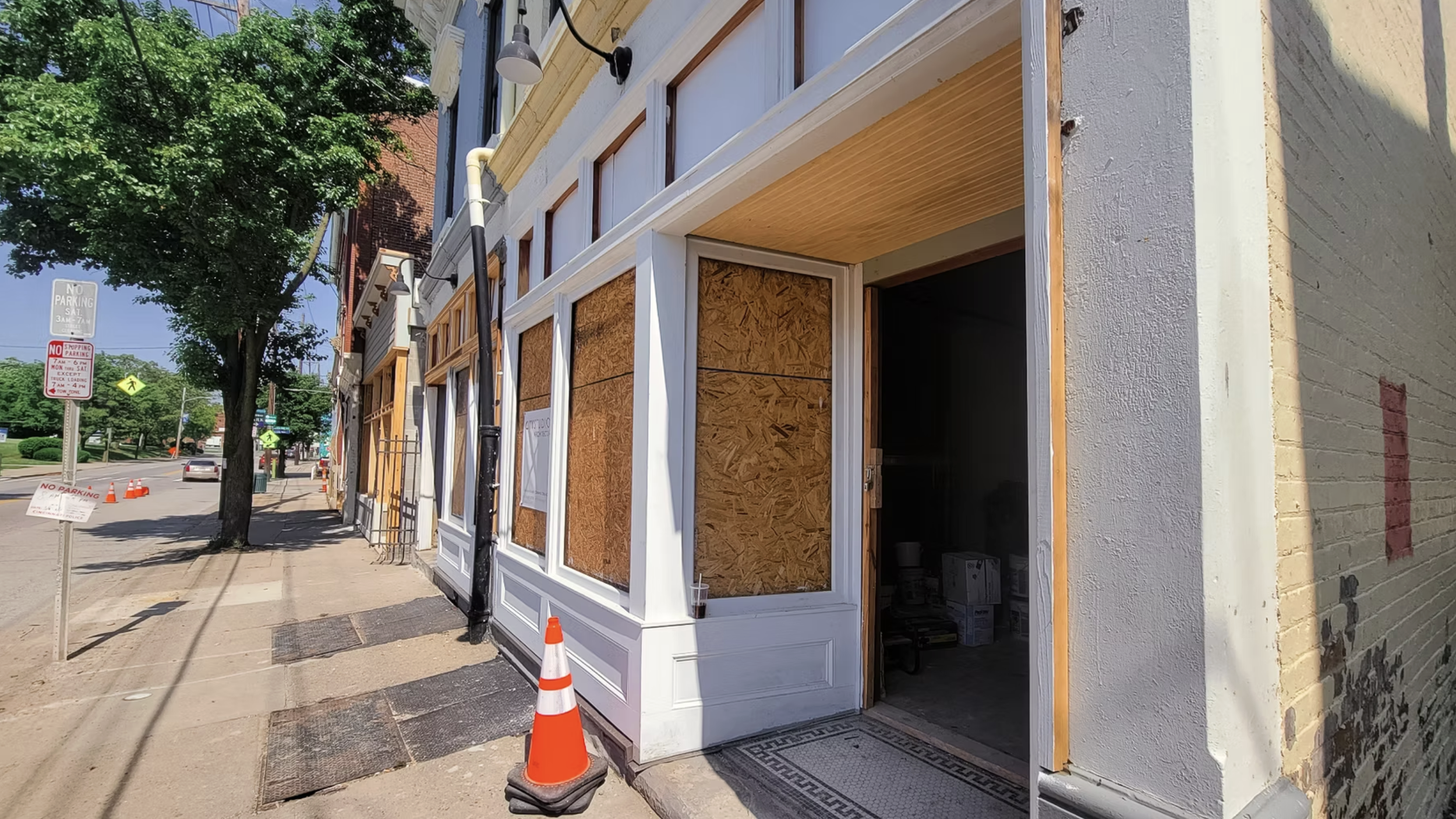 A building under construction with boarded-up windows and an open doorway, located on a sidewalk next to a tree and street with parking signs and traffic cones.