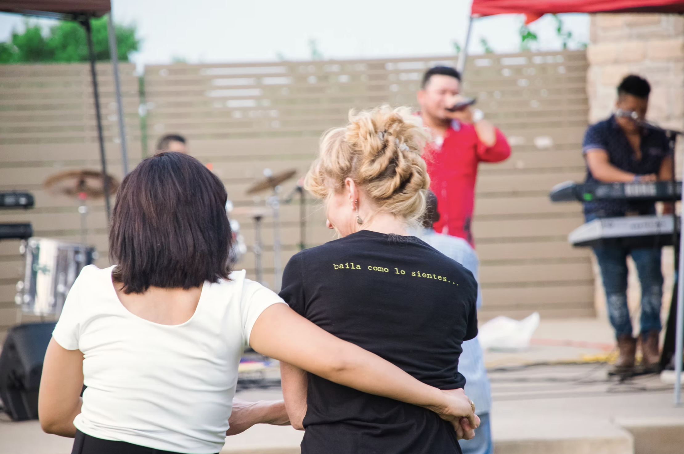 Two women dancing closely at an outdoor music event, with a band playing on stage behind them.