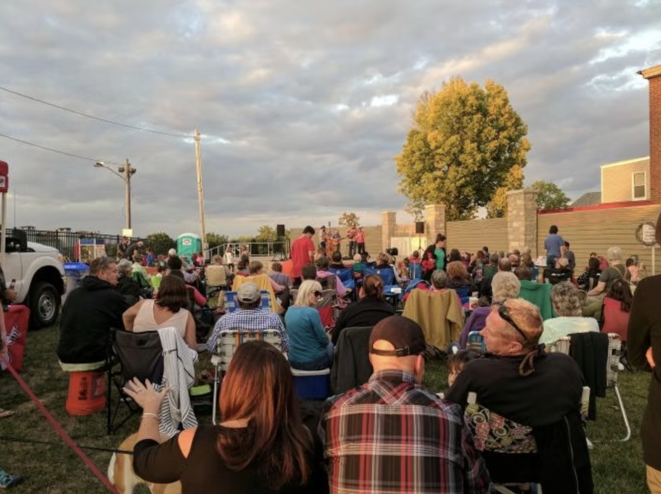 An outdoor event with a crowd of people seated in chairs watching a performance or speech on a stage. The scene is set in the evening with cloudy sky and some trees in the background.