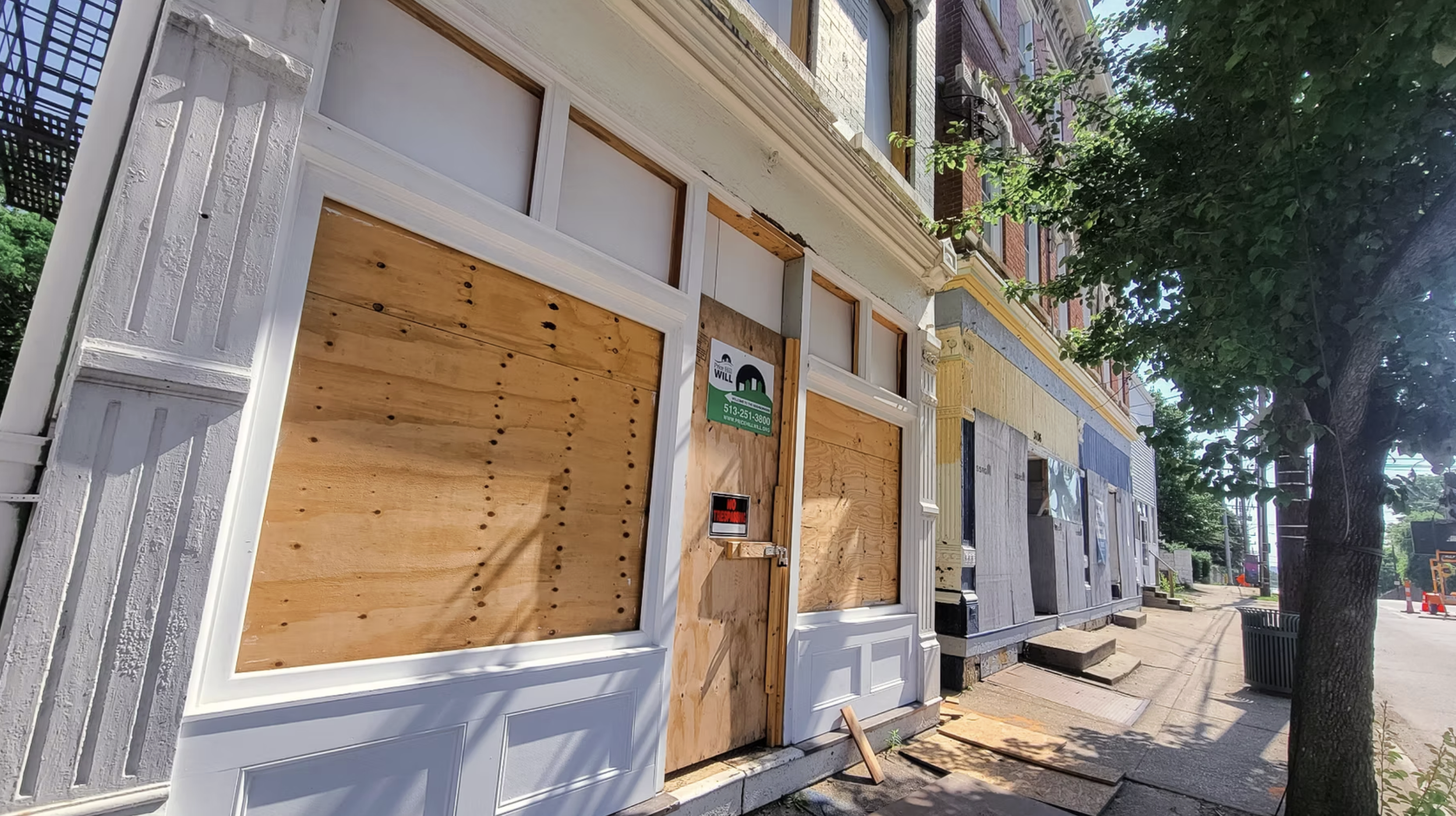 Row of buildings under renovation with boarded-up windows and doors, surrounded by trees on a sidewalk, with some construction signs and materials visible.