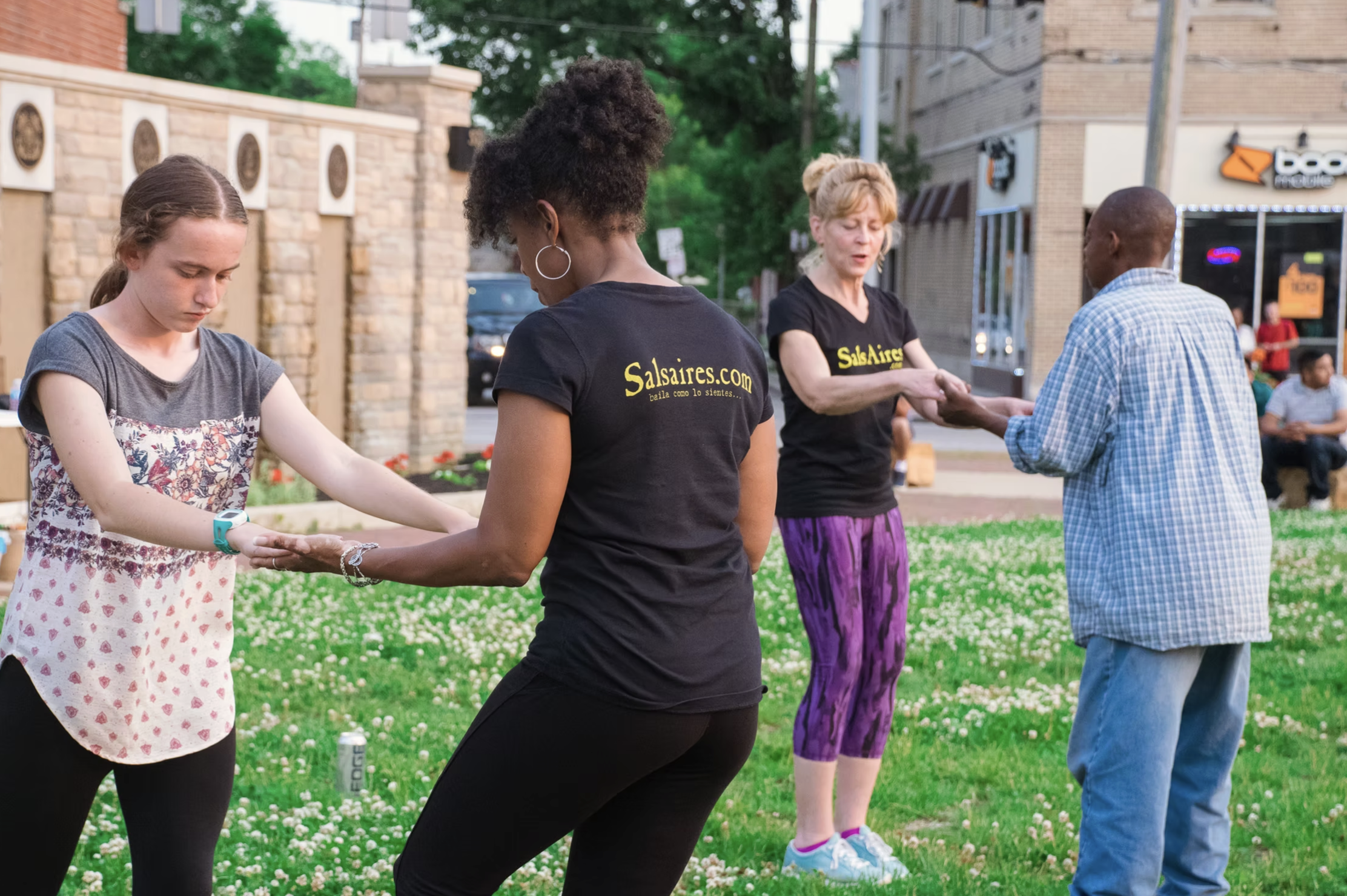 Four people dancing in a circle outdoors on a grassy area with clovers, with a brick wall and buildings in the background. Two women are wearing black T-shirts with 'Salsares.com' printed on them.