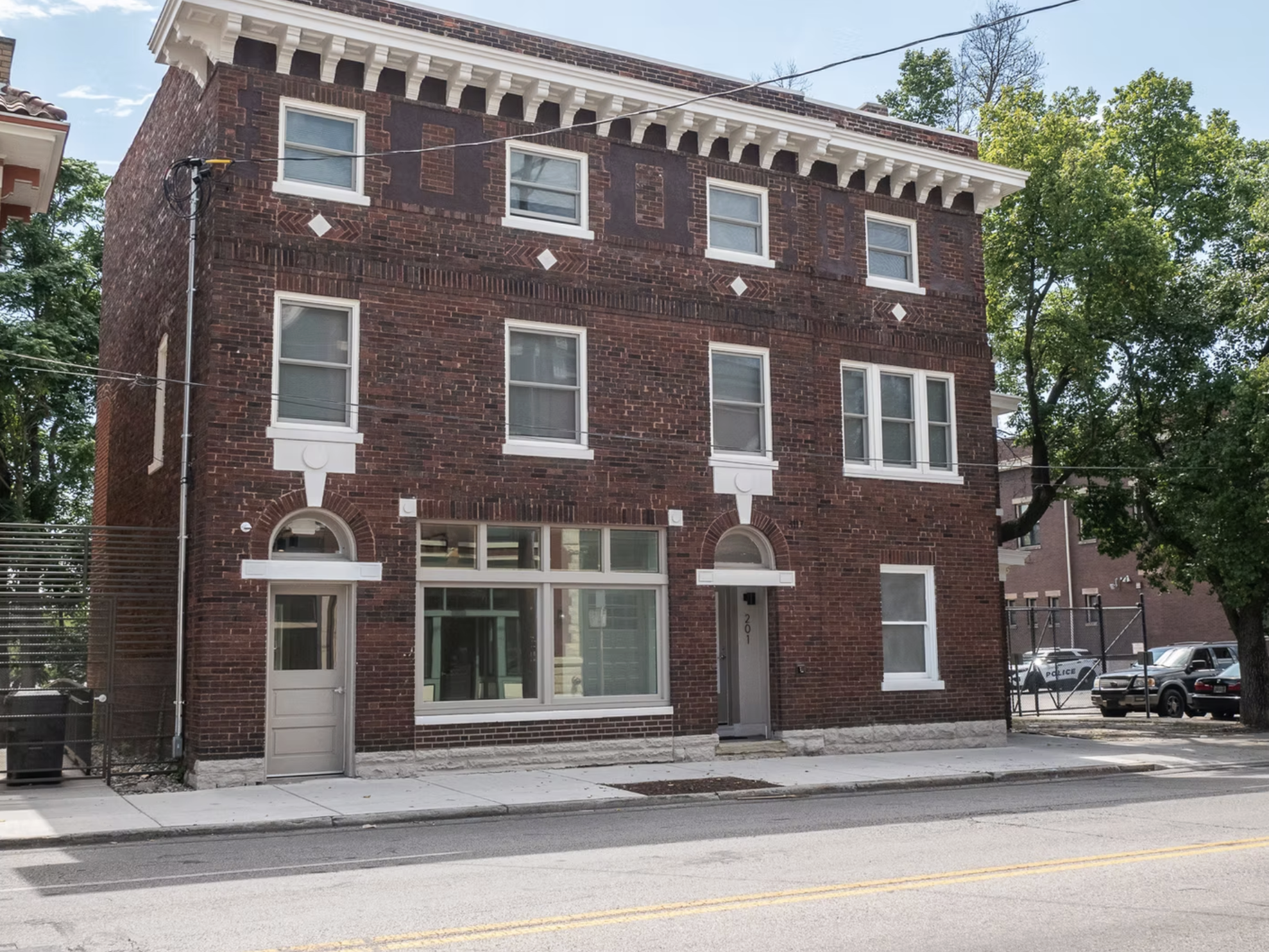 A three-story red brick building with decorative white accents and a flat roof, located on a city street with trees and parked cars visible in the background.