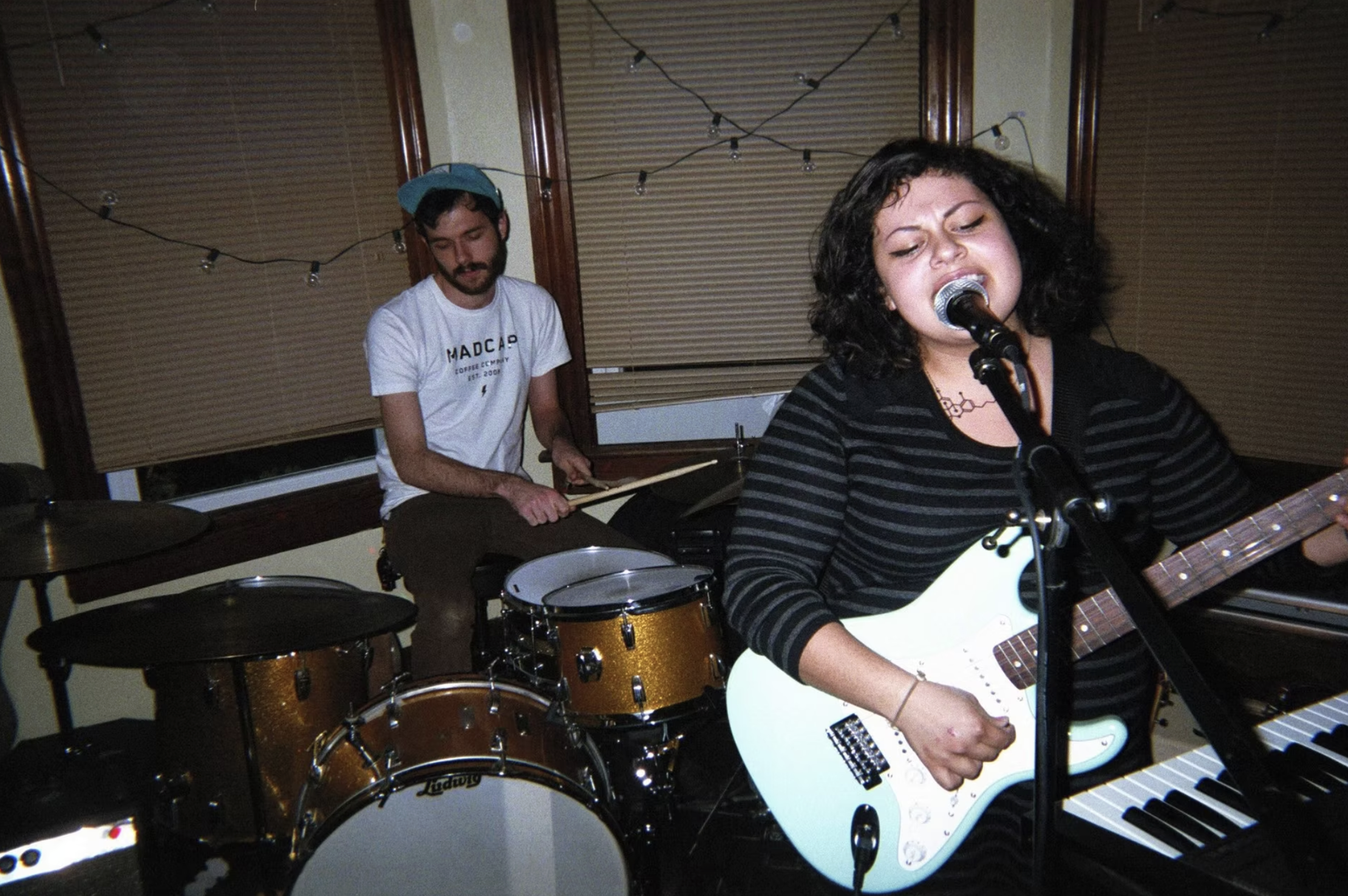 A woman singing and playing guitar and keyboard, with a man in the background playing drums, performing in a room with wood-paneled windows and string lights.