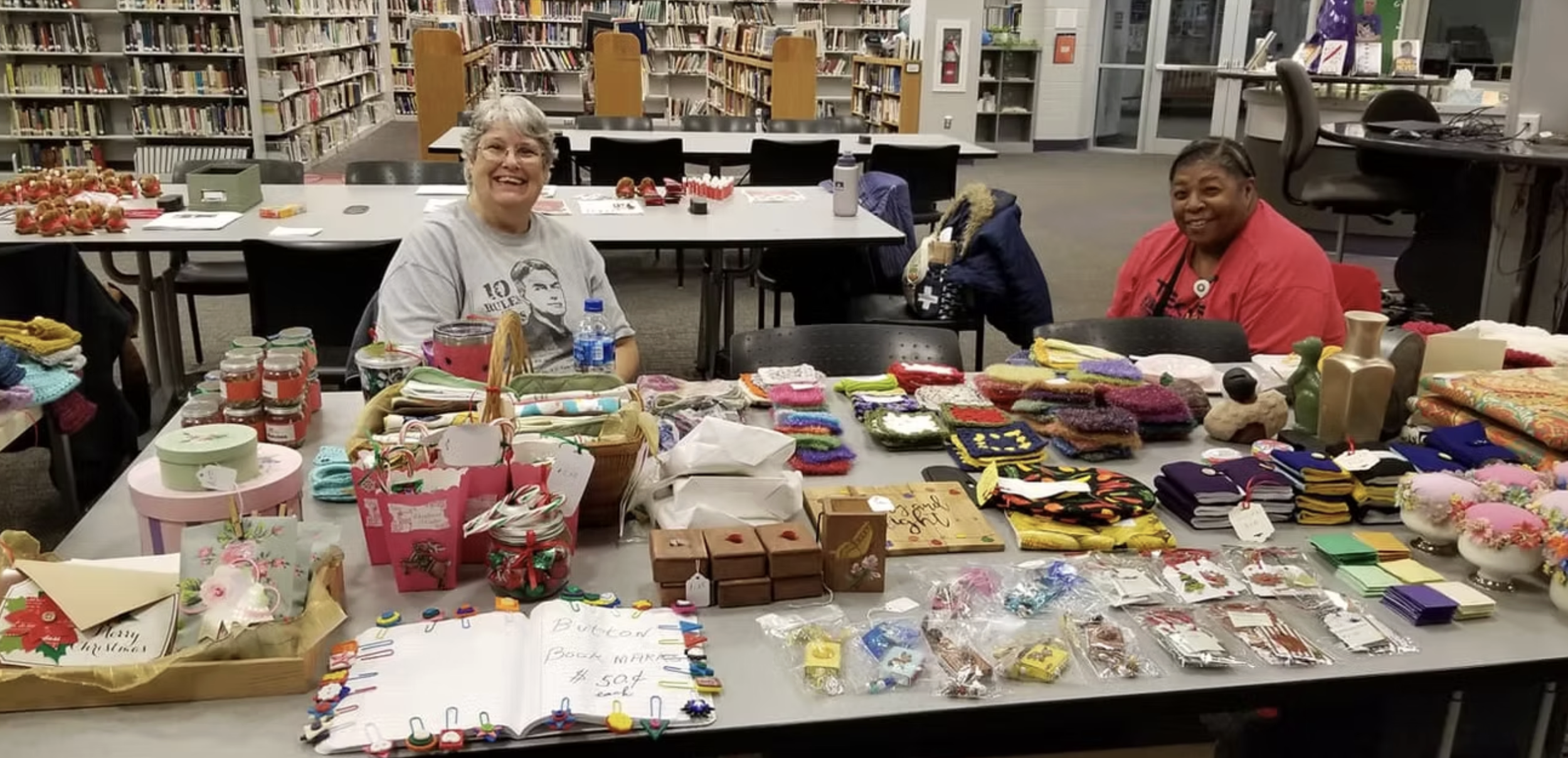 Two women smiling behind a table filled with holiday crafts and decorations at a library or community center.