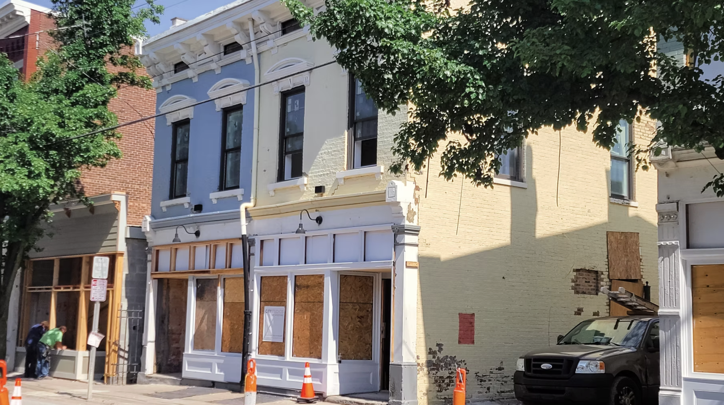 Street view of a row of buildings with storefronts undergoing renovation; boarded-up windows and doors, construction cones on the sidewalk, and two workers in green shirts inspecting a storefront.