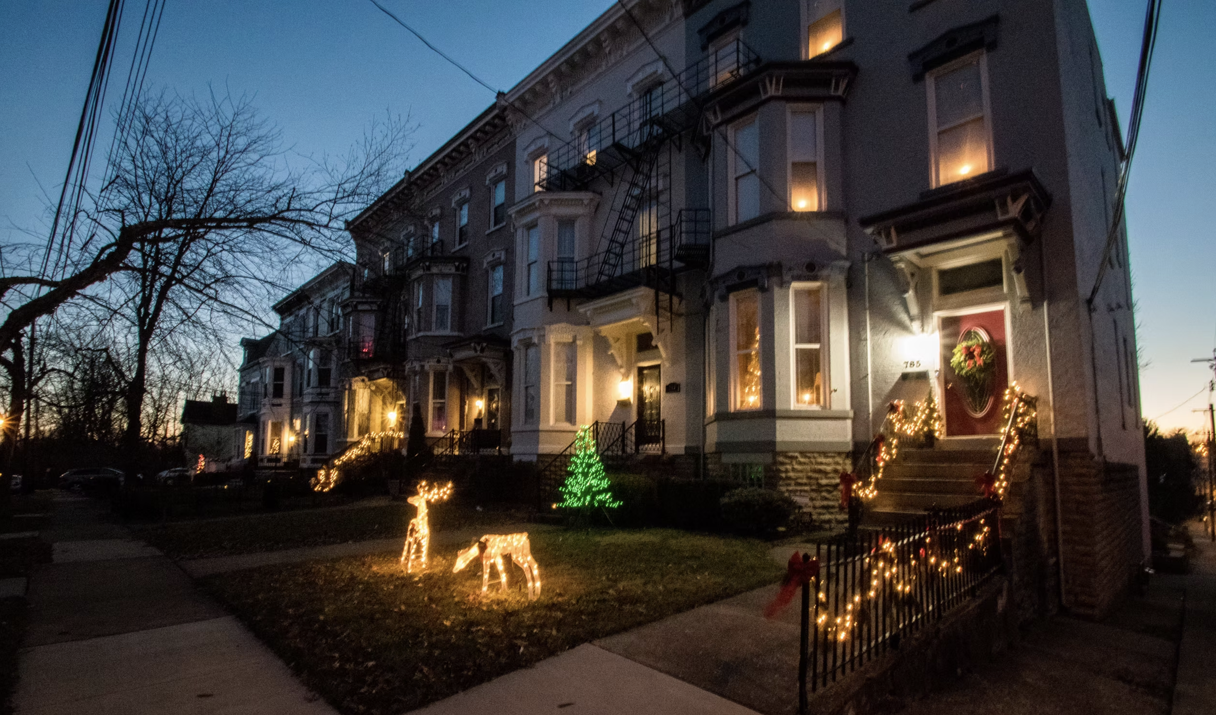 A row of Victorian-style houses decorated with Christmas lights, including a green light-up Christmas tree and illuminated reindeer figures on the lawn. The houses are lit from within, and the steps leading up to the doors are decorated with strings 
