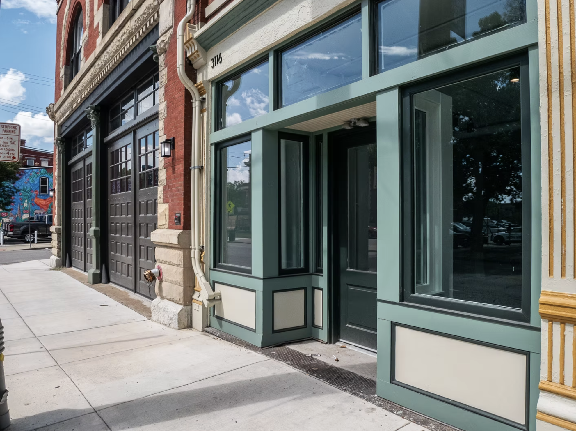 Street view of storefront with large windows and black door, green trim, adjacent to brick building with garage door, sidewalk in foreground, mural visible in background at corner of street