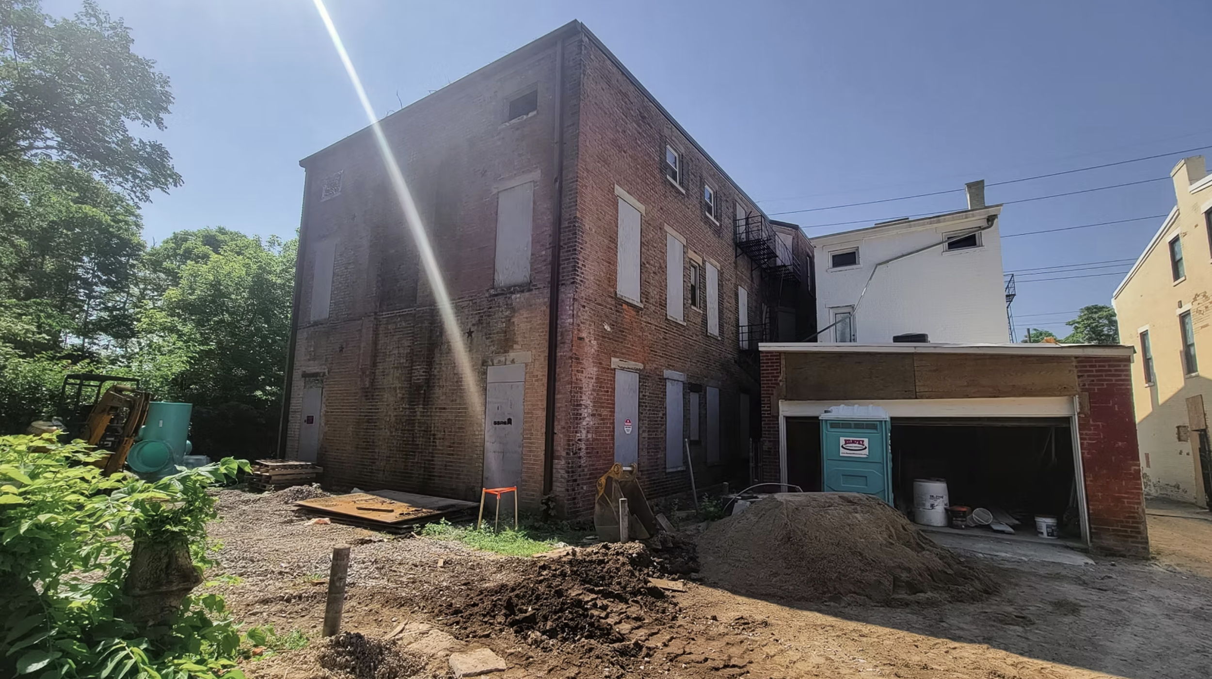 Construction site with a three-story brick building, boarded windows, green trees on the left, a pile of dirt, construction materials, and portable toilets in the foreground.