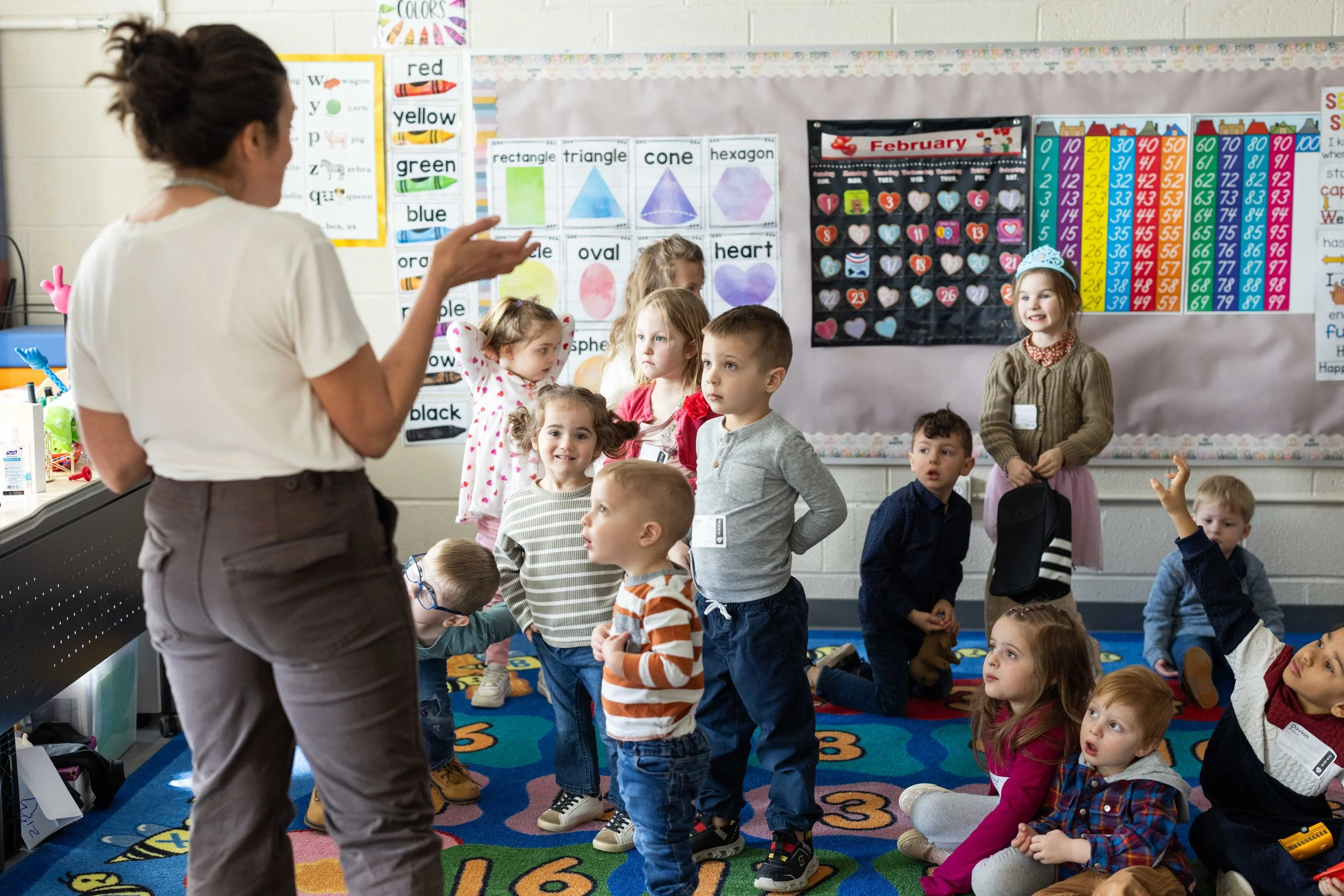 A teacher is speaking to a group of young children in a classroom. The children are standing or sitting on a colorful alphabet-themed rug, listening attentively. The classroom has educational posters on the wall, including shapes, colors, and a calendar.