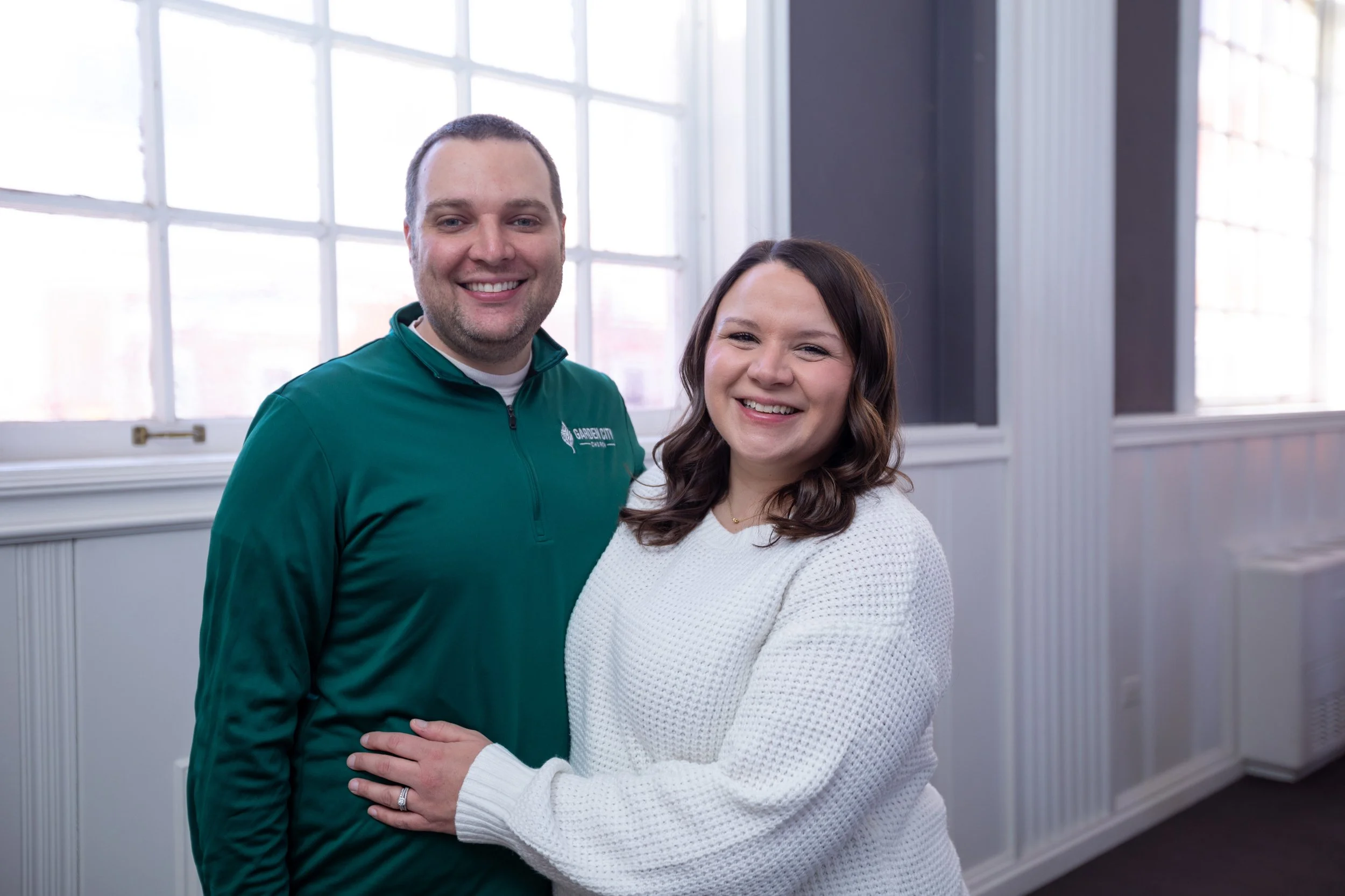 A smiling couple standing close together indoors near large windows with white frames, the man wearing a green jacket with a logo and the woman in a white sweater.