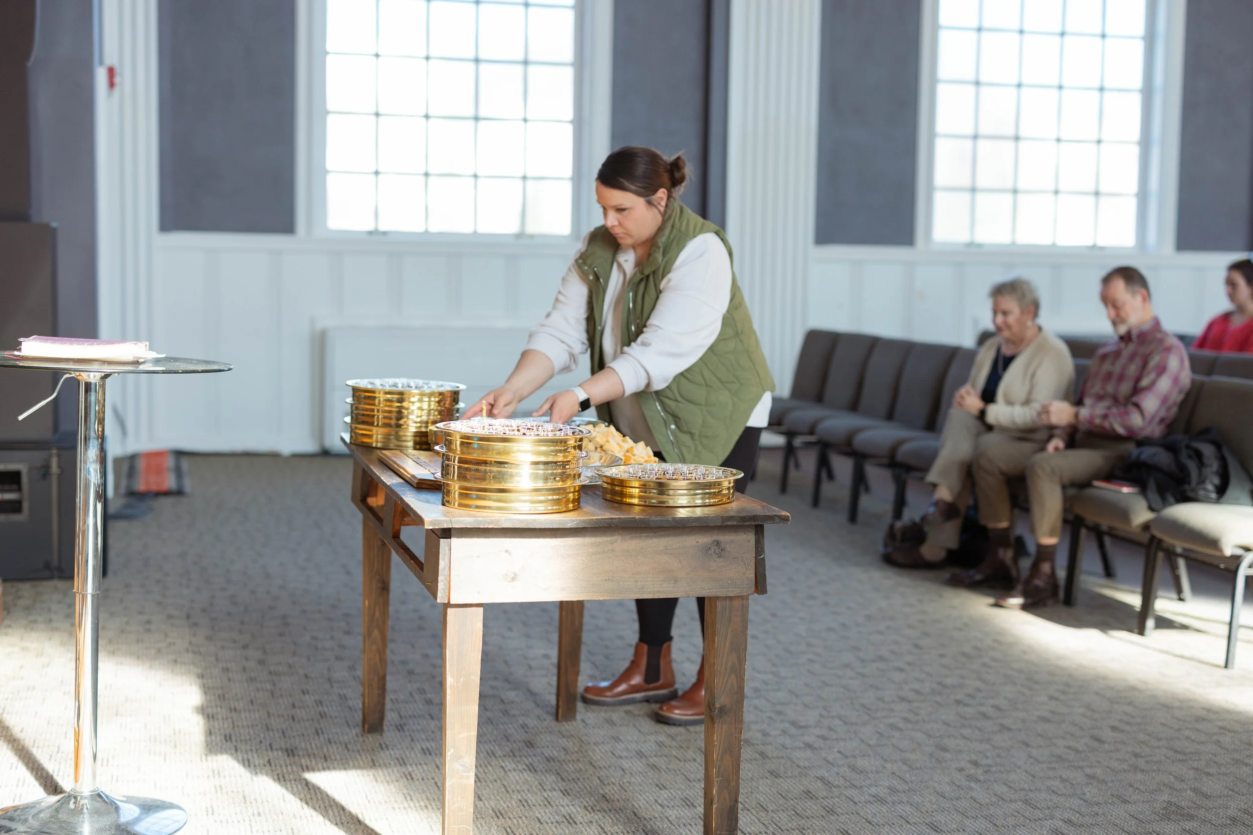 Woman arranging trays of small desserts on a table in a room with metal chairs and large windows.