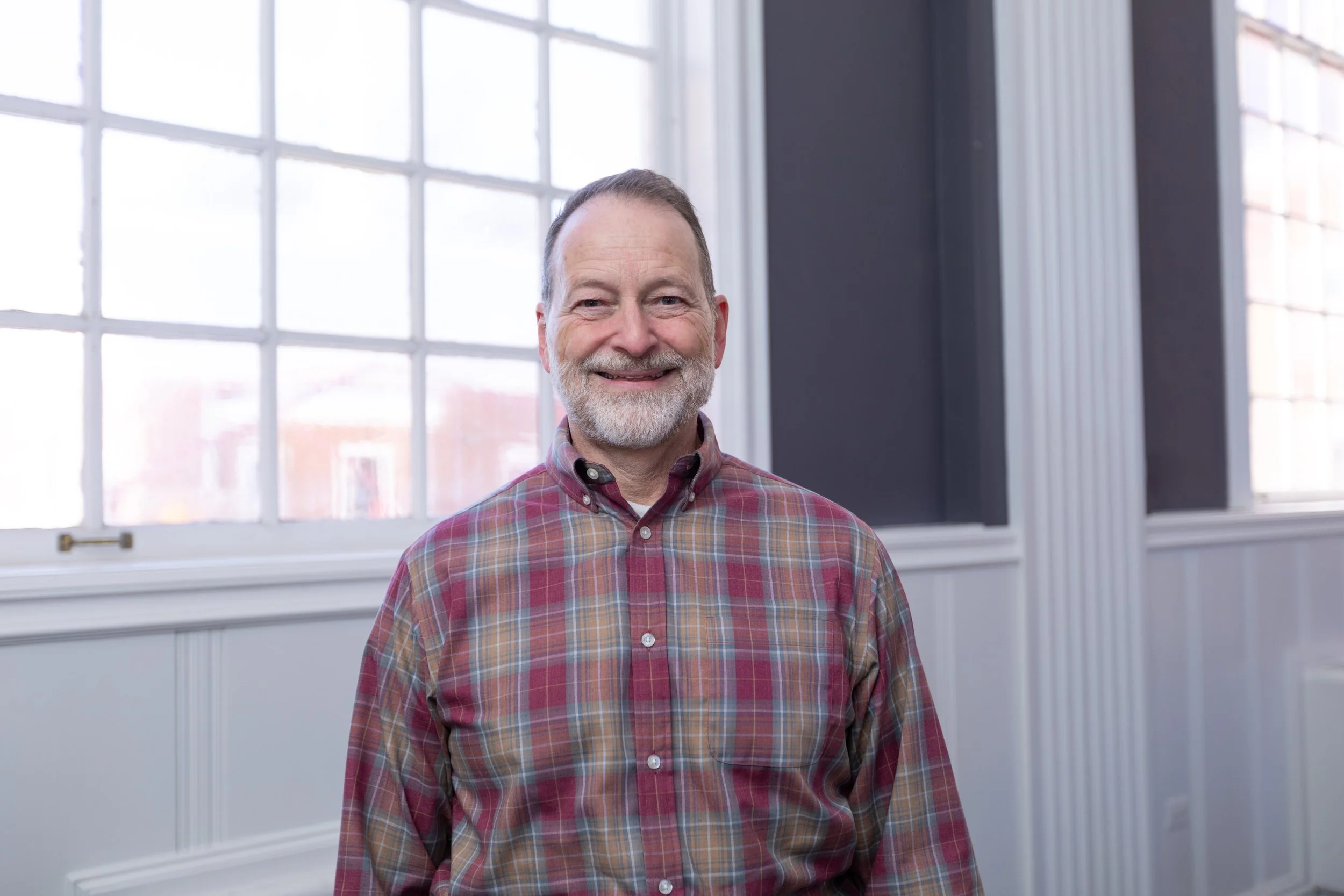 An older man with a beard and mustache smiling, wearing a red plaid shirt, standing in front of large windows with white frames in a bright room.