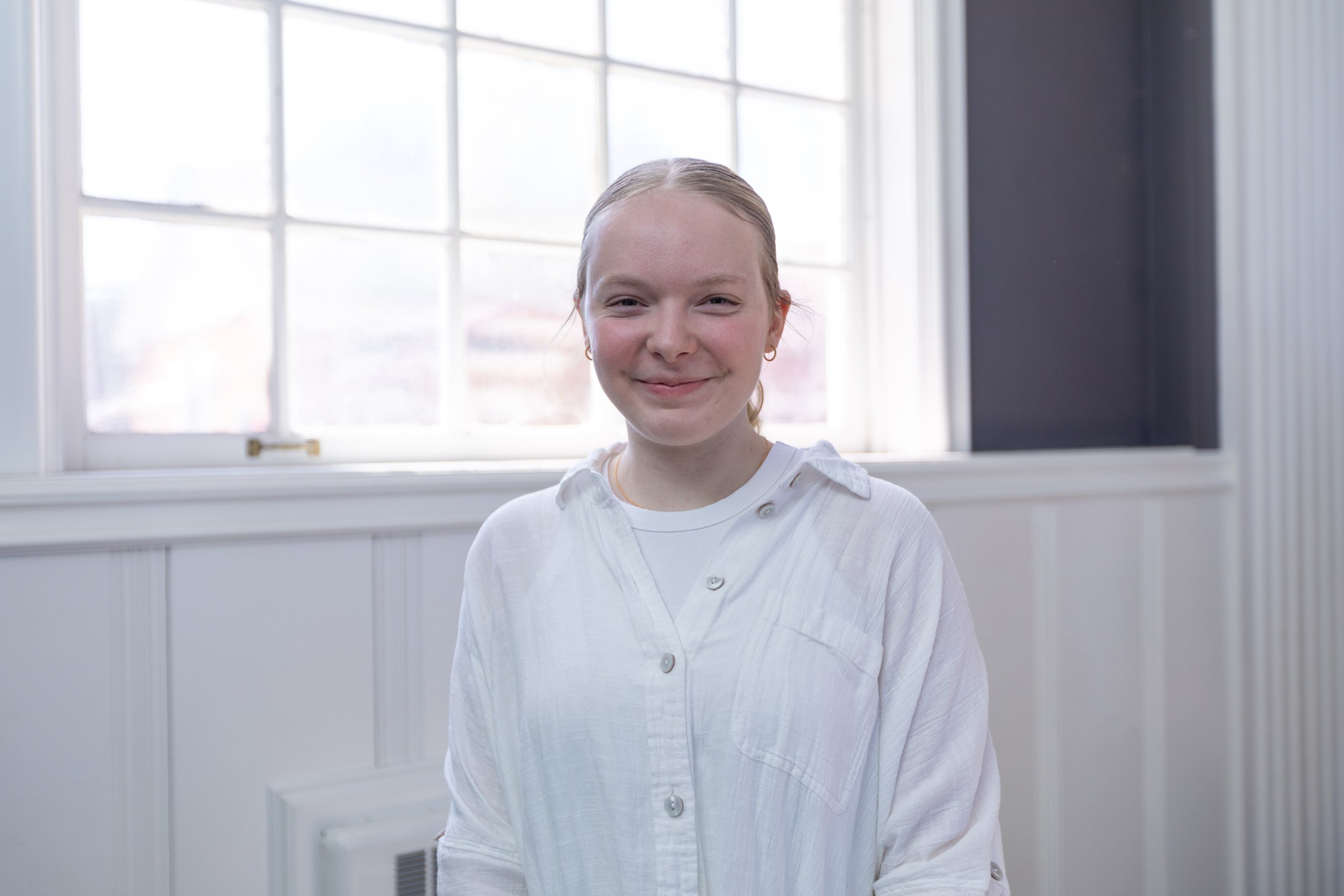 A young woman with blonde hair tied back, wearing a white shirt, standing indoors in front of a large window with white panes and a dark wall on the right side.