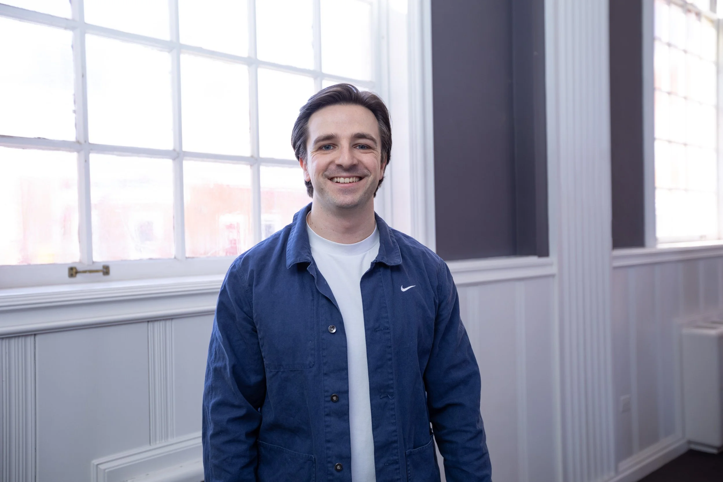A smiling young man with dark hair, wearing a blue jacket over a white shirt, standing inside a room with large windows and white paneling.