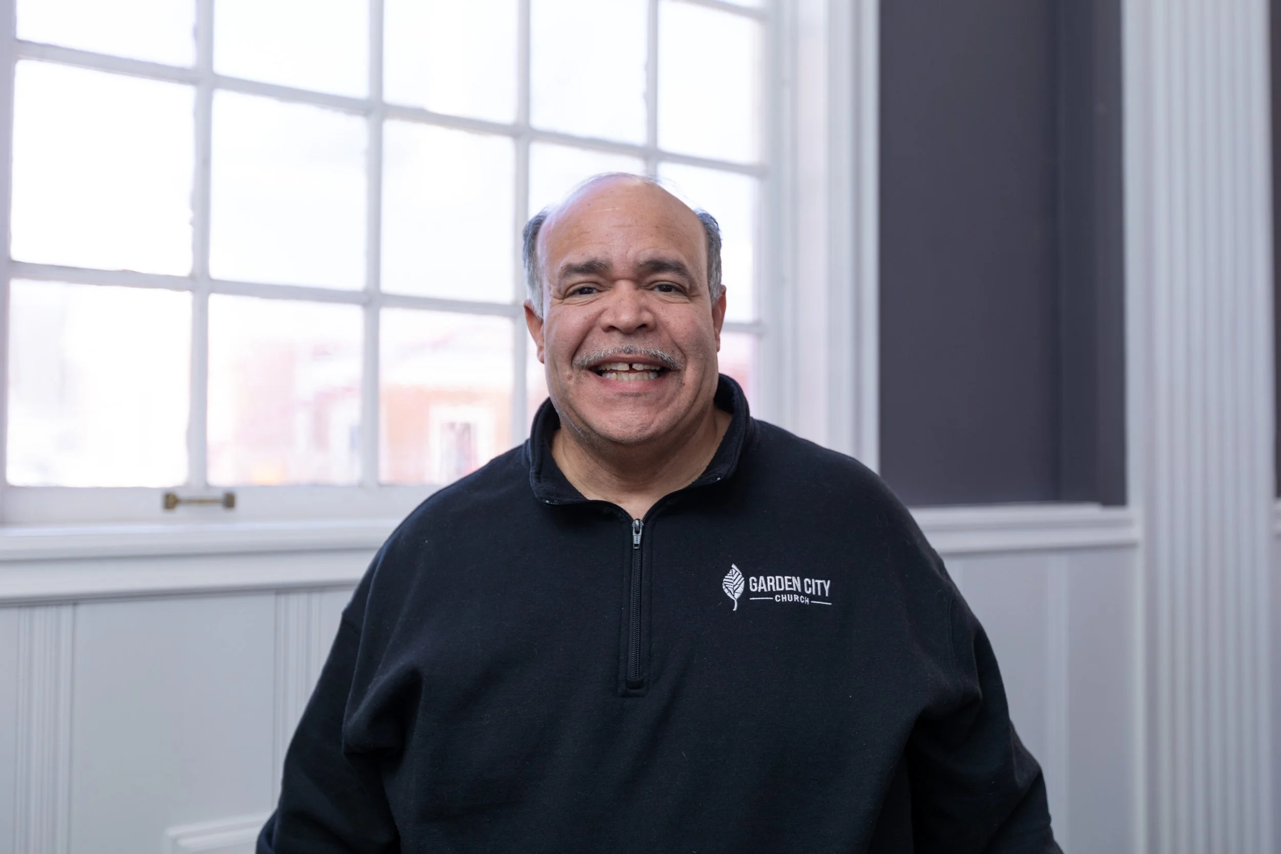 A smiling middle-aged man wearing a black jacket with the logo 'Garden City Church' standing indoors in front of a large window with multiple panes.