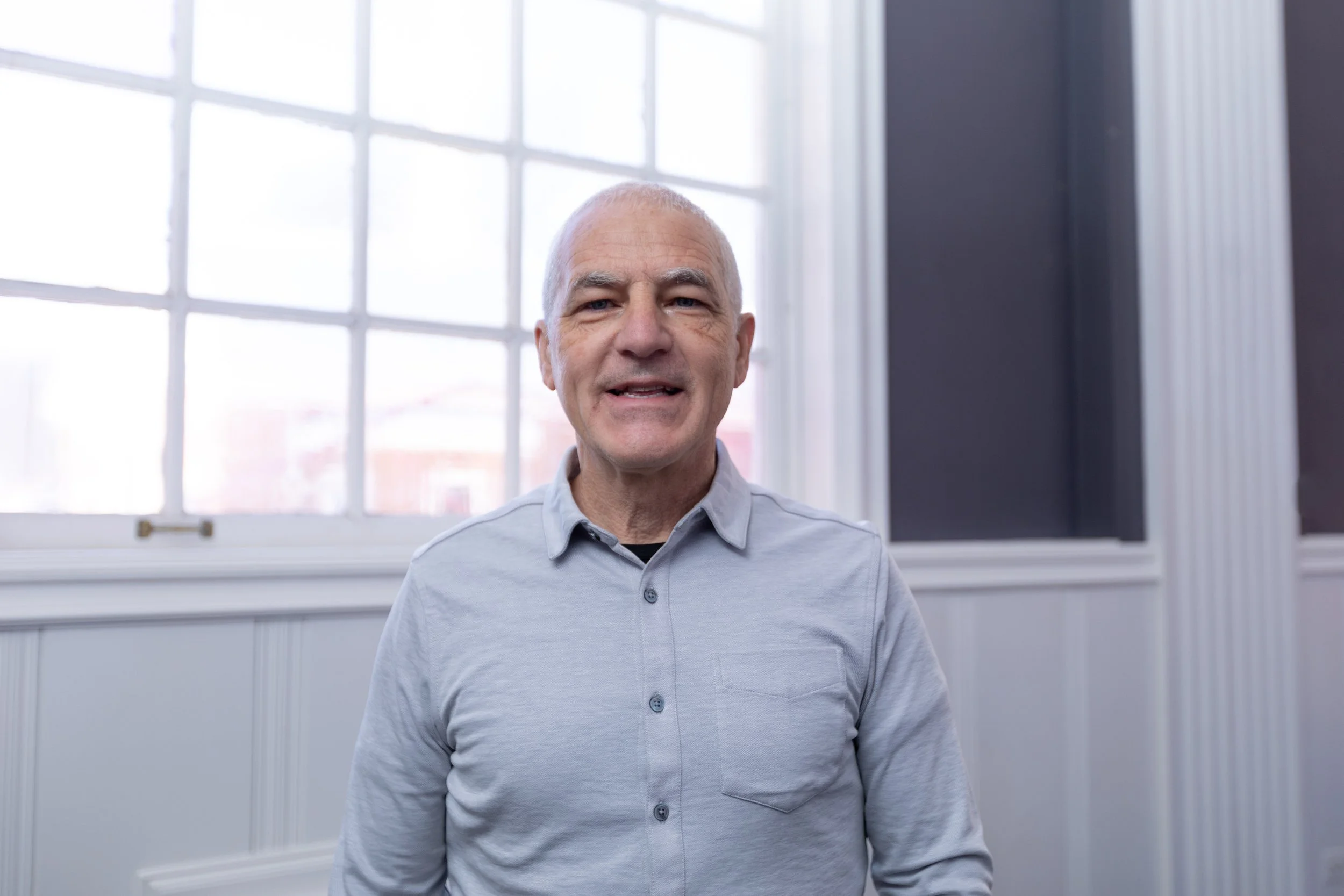 A middle-aged man with short gray hair and wearing a light gray button-up shirt, standing indoors in front of a large window with white framing. Soft natural light bleeds into the room.