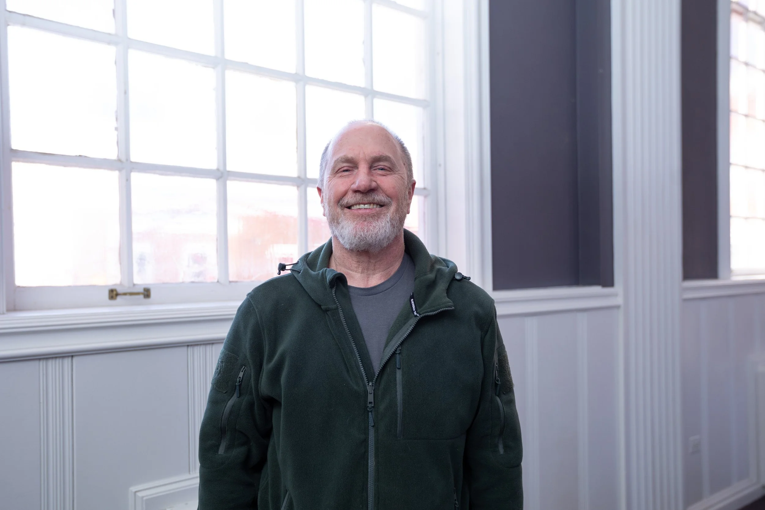 A smiling elderly man with a beard standing indoors near large windows, dressed in a dark jacket and gray shirt.