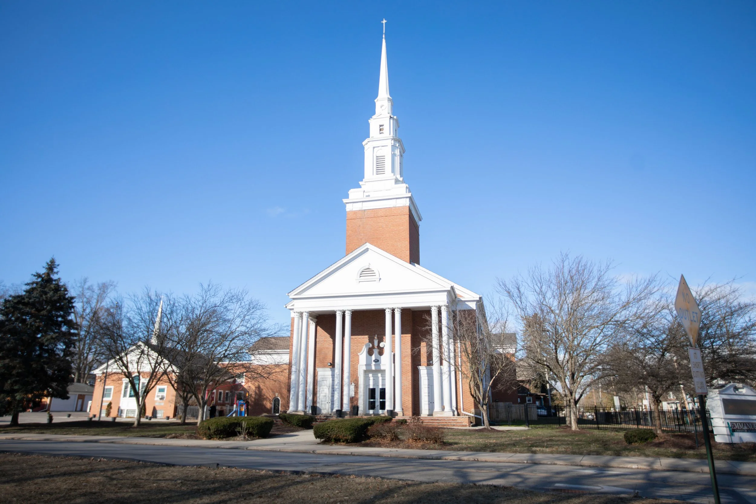 A church building with a tall white steeple topped with a cross, white columns at the entrance, and a brick facade, set against a clear blue sky with trees and residential buildings in the background.