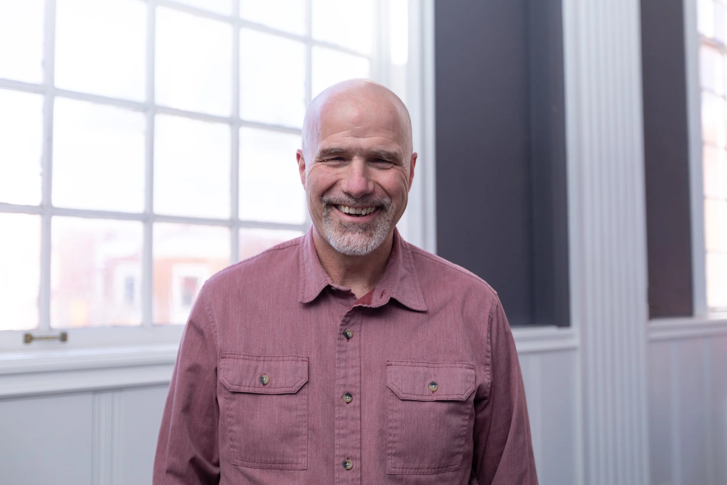 A smiling bald man with gray facial hair wearing a pink button-up shirt standing in front of a large window with multiple panes, inside a modern building.