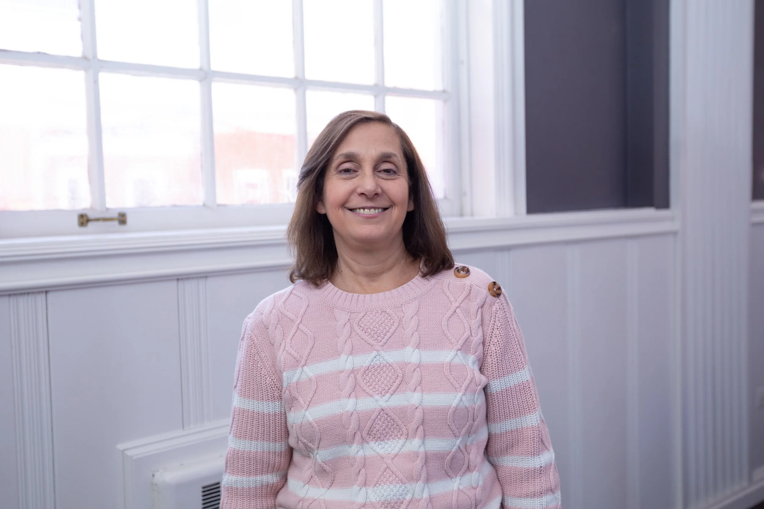A middle-aged woman with shoulder-length brown hair, smiling in front of a large window with white framing and interior white wall paneling, wearing a pink sweater with decorative buttons on the shoulder.