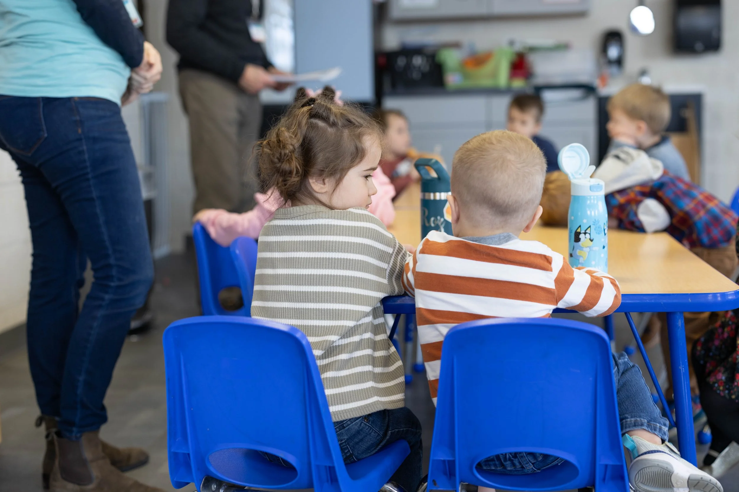 Children sitting at blue chairs around a table, with two adults standing nearby in a classroom or daycare setting.