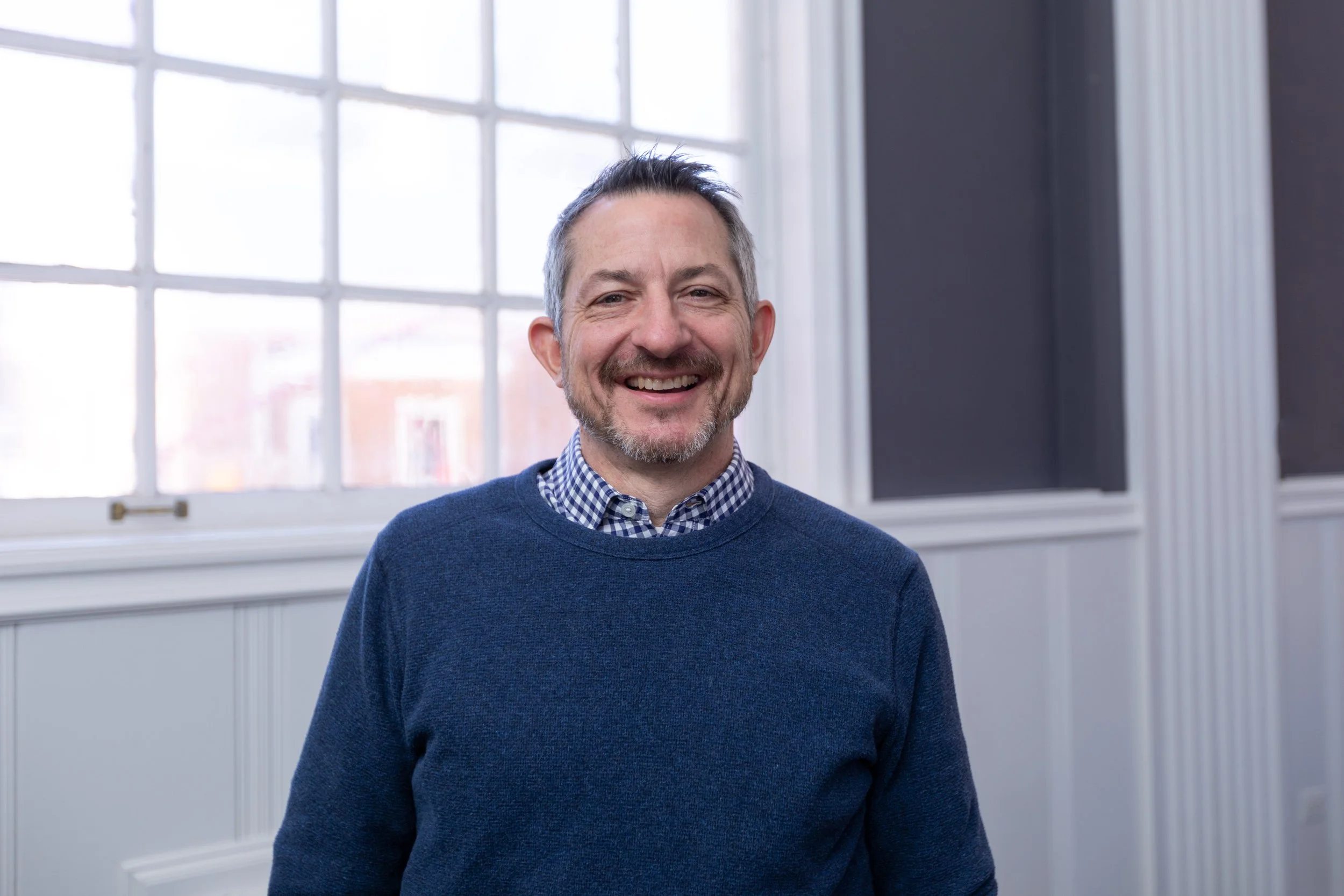 A middle-aged man with short, graying hair and a beard, smiling, wearing a blue sweater over a checkered shirt, standing inside a room with large windows and white walls.