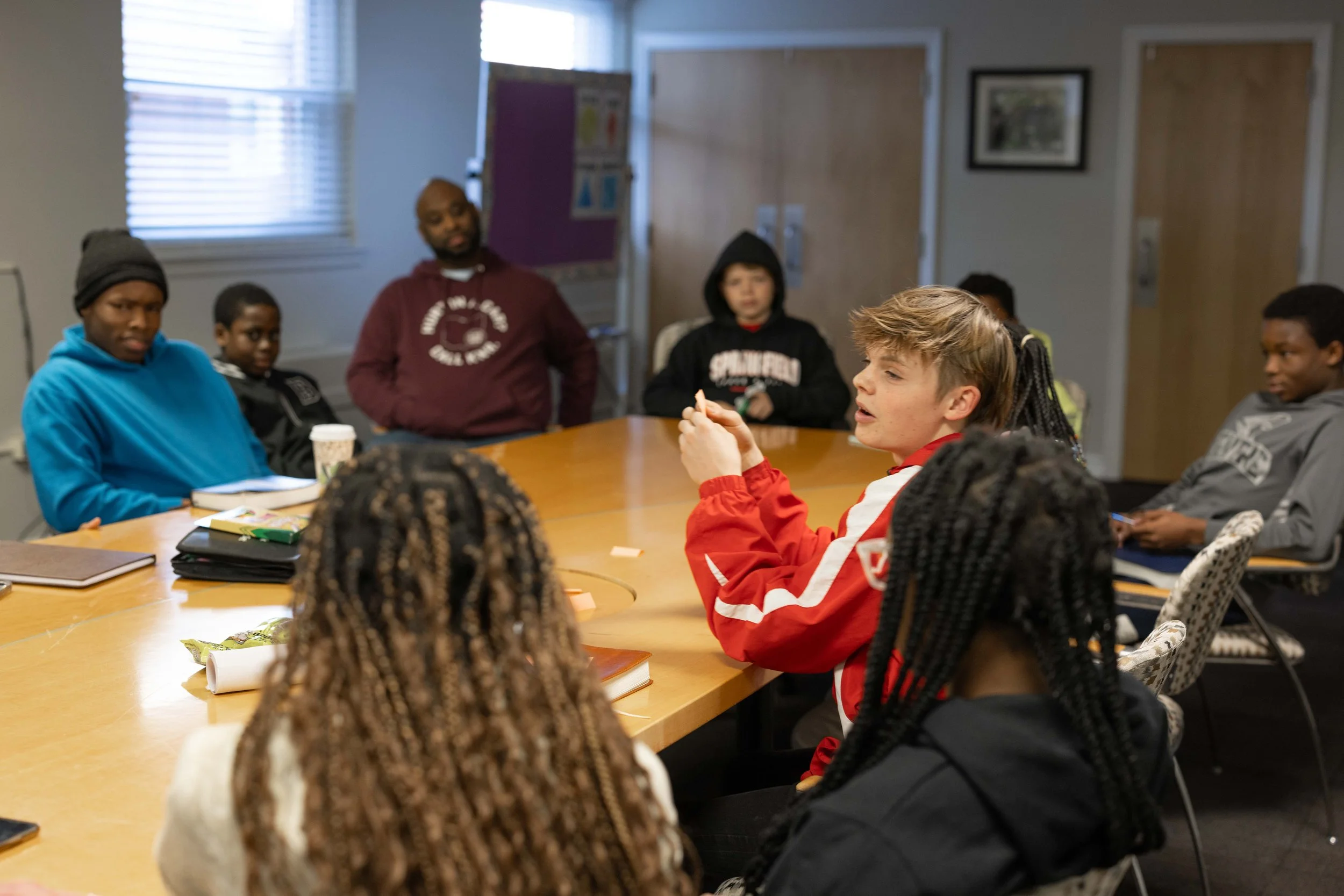 Group of diverse children sitting around a large oval table in a classroom during a discussion or activity, with notebooks and snacks on the table, and a man standing at the back.