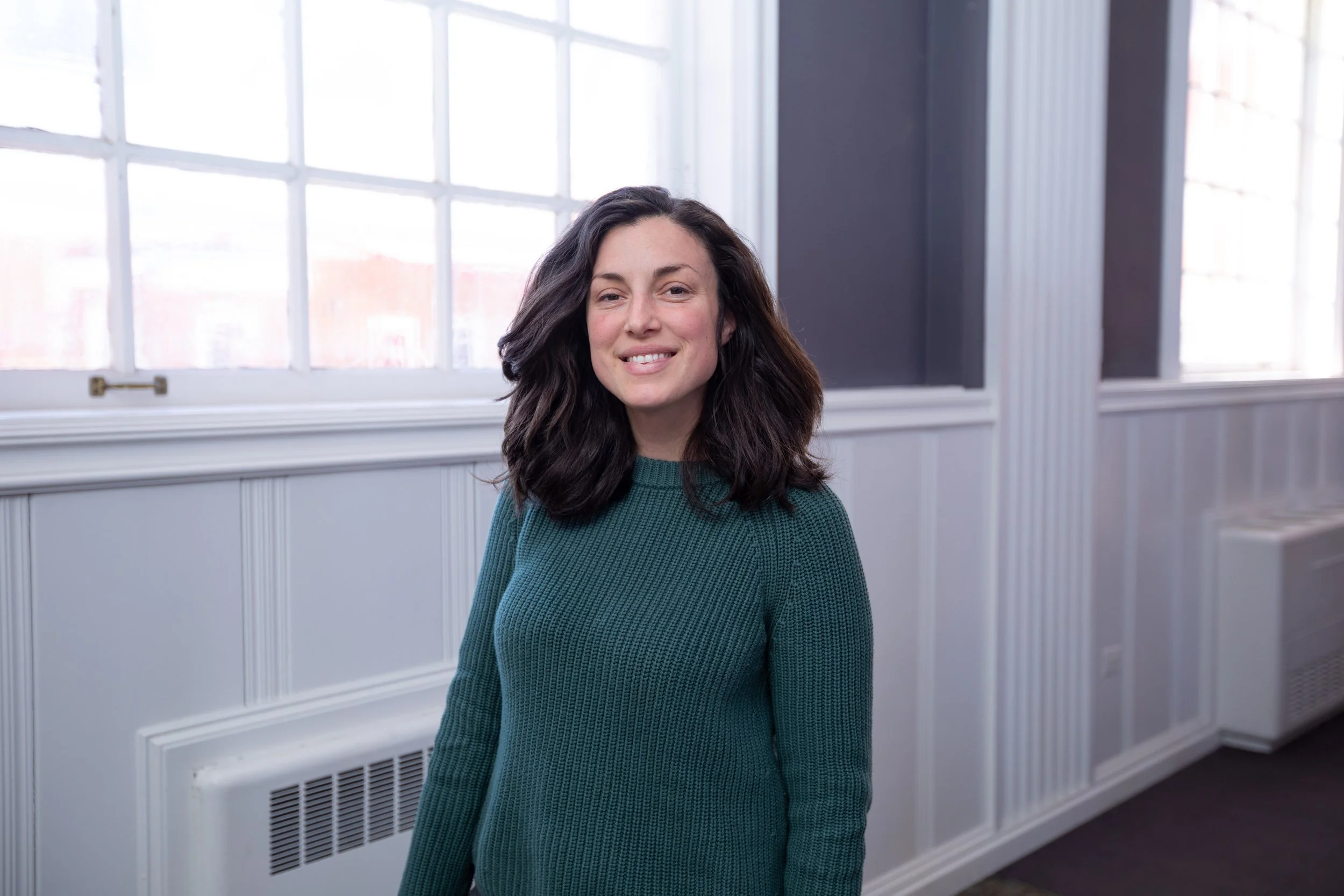 A woman with long dark hair smiling, wearing a teal sweater, standing in front of large windows with white frames in a room with white wall paneling.