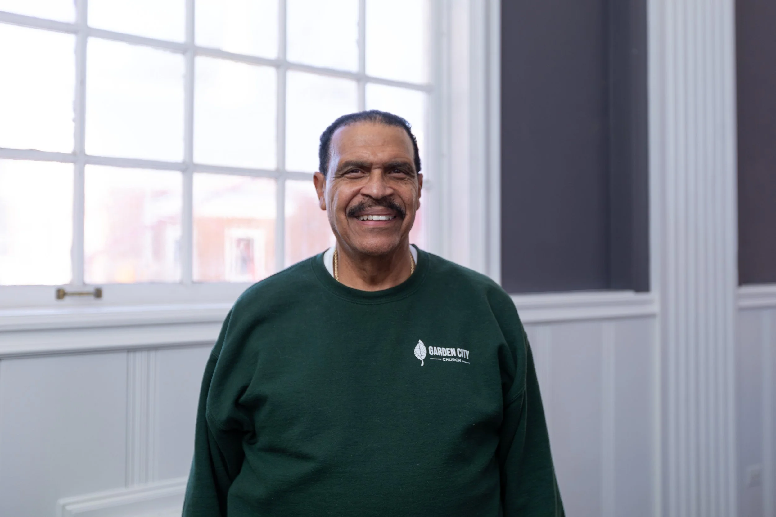 A smiling man with a mustache and dark hair, wearing a green sweatshirt with 'Garden City Church' logo, standing in front of a large window with multiple panes.
