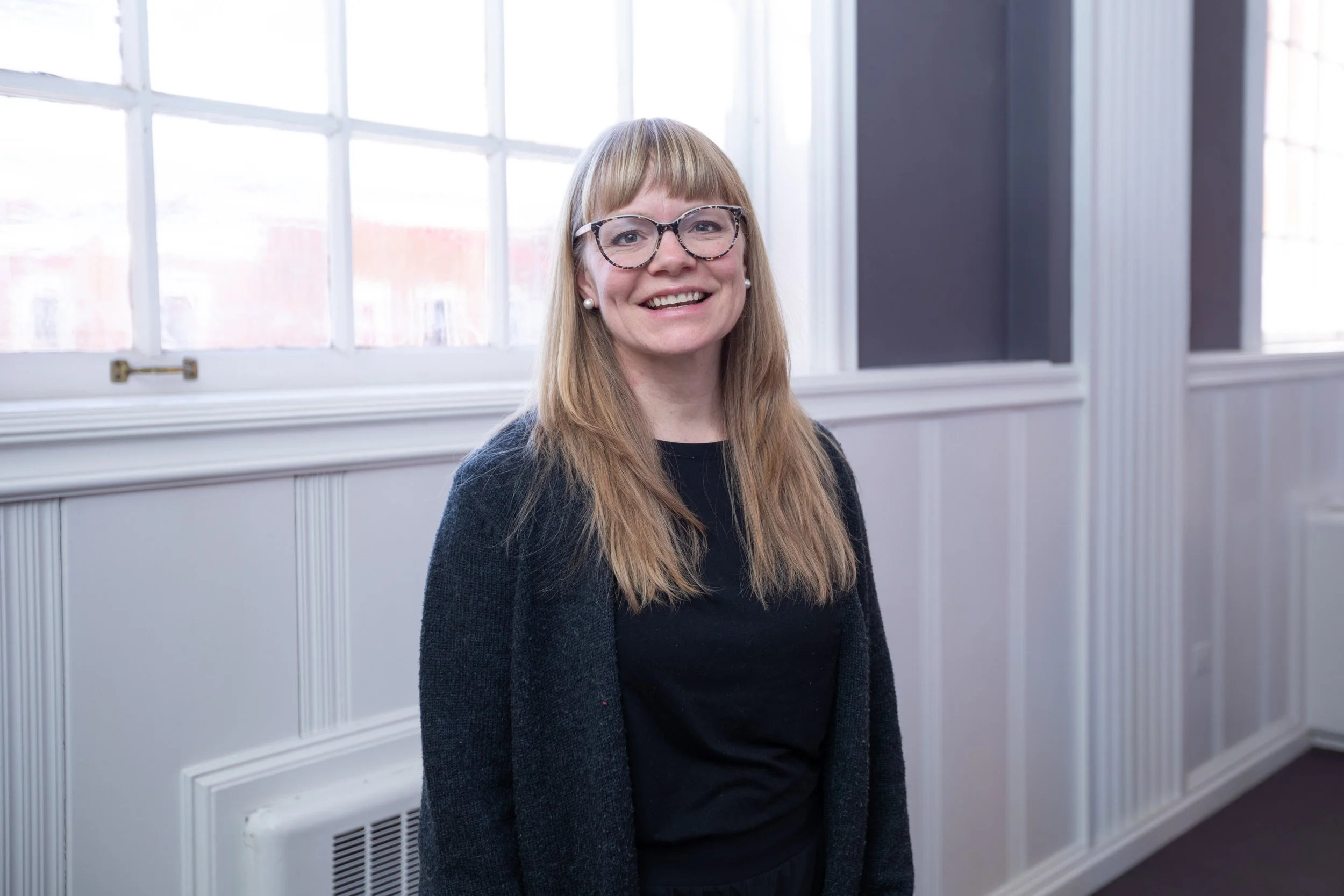 A woman with long blonde hair, wearing glasses with tortoiseshell frames, pearl earrings, a black top, and a dark cardigan, smiling while standing indoors near a large window with a white frame.