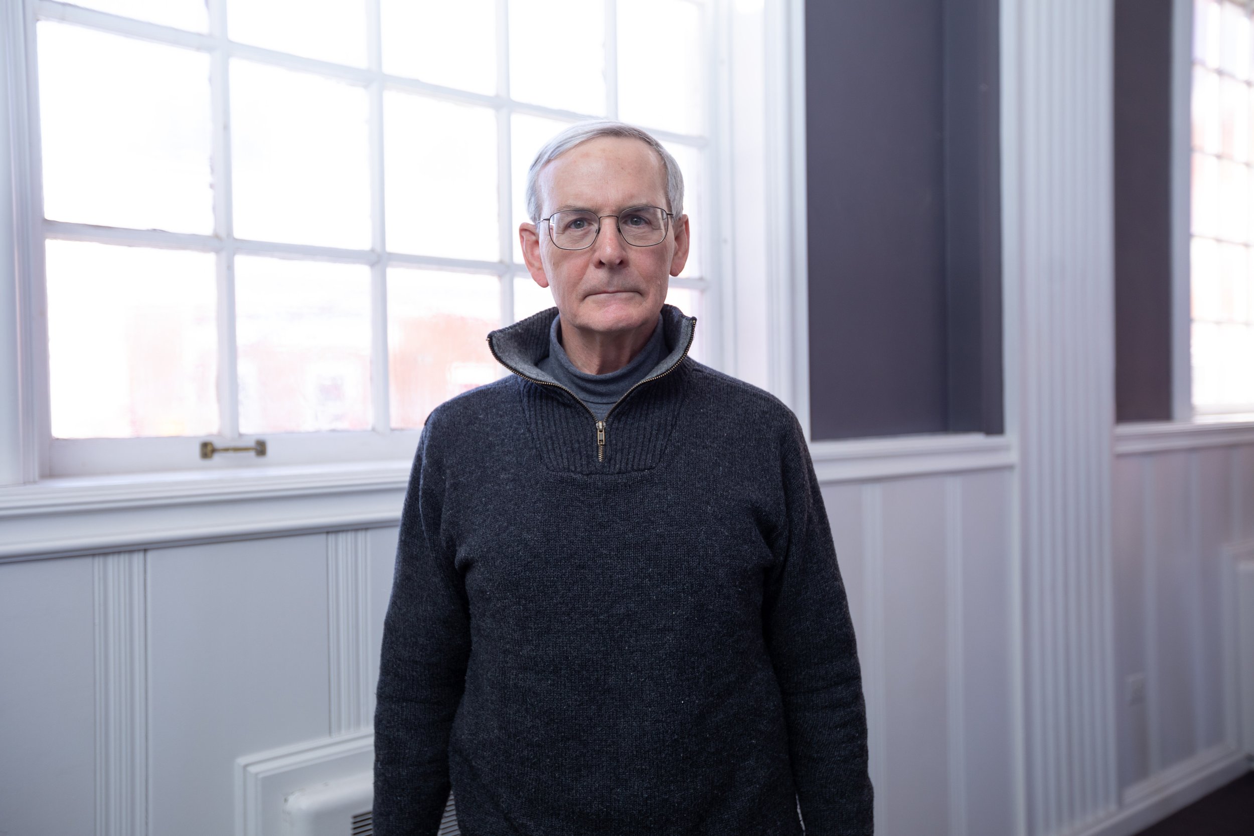 Older man wearing glasses and a dark sweater, standing in front of large windows in a bright room.
