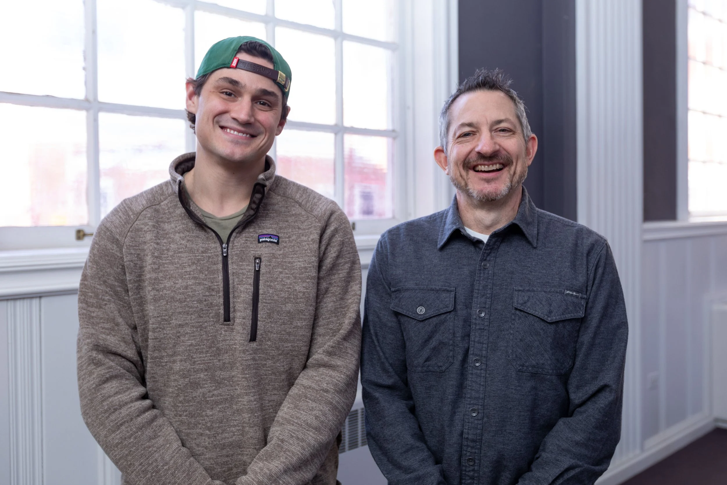 Two men standing indoors in front of large windows, smiling at the camera. One man is wearing a brown sweater and a green cap backwards, while the other man is in a dark gray button-up shirt.