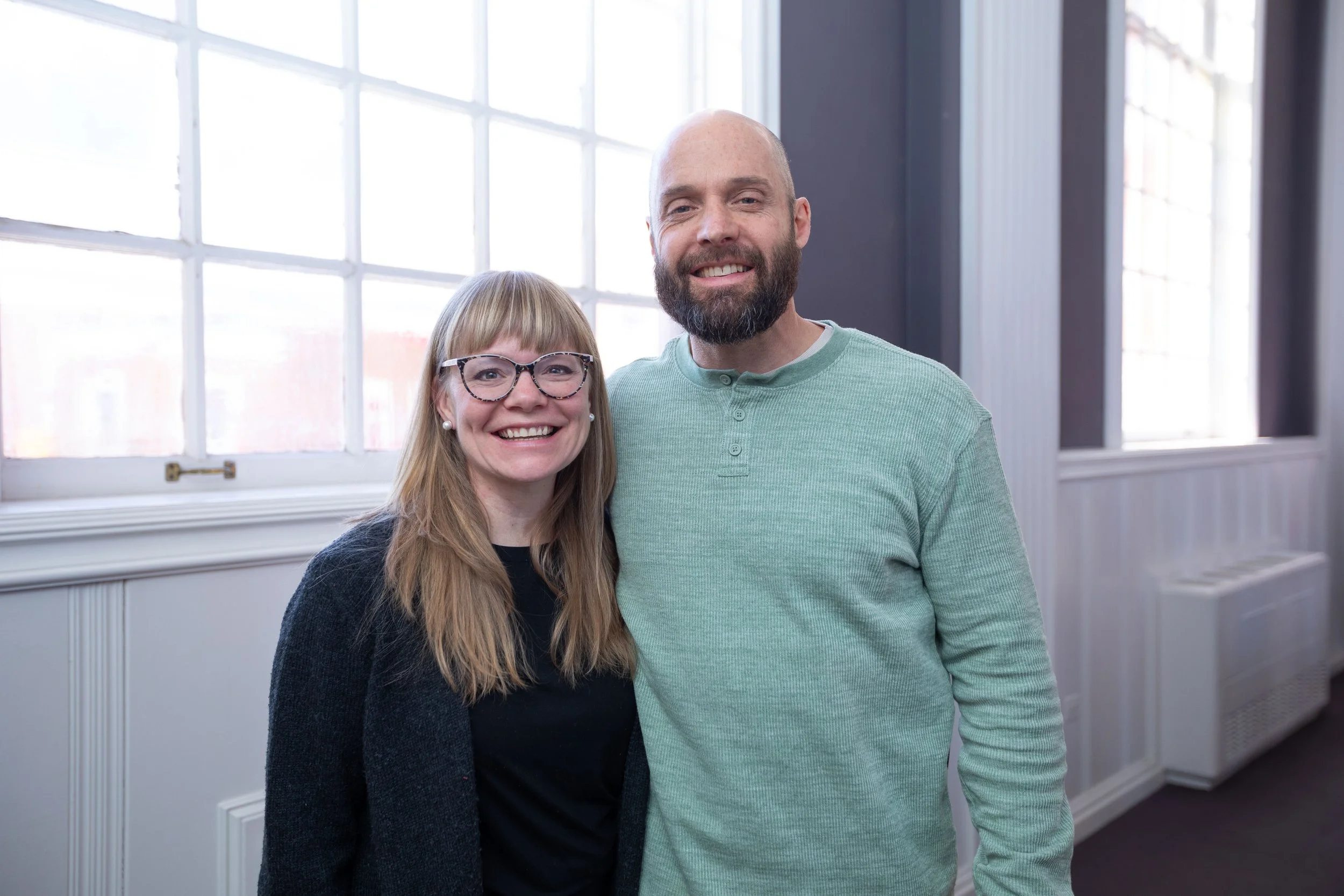 A man and woman smiling and posing together indoors near large windows.