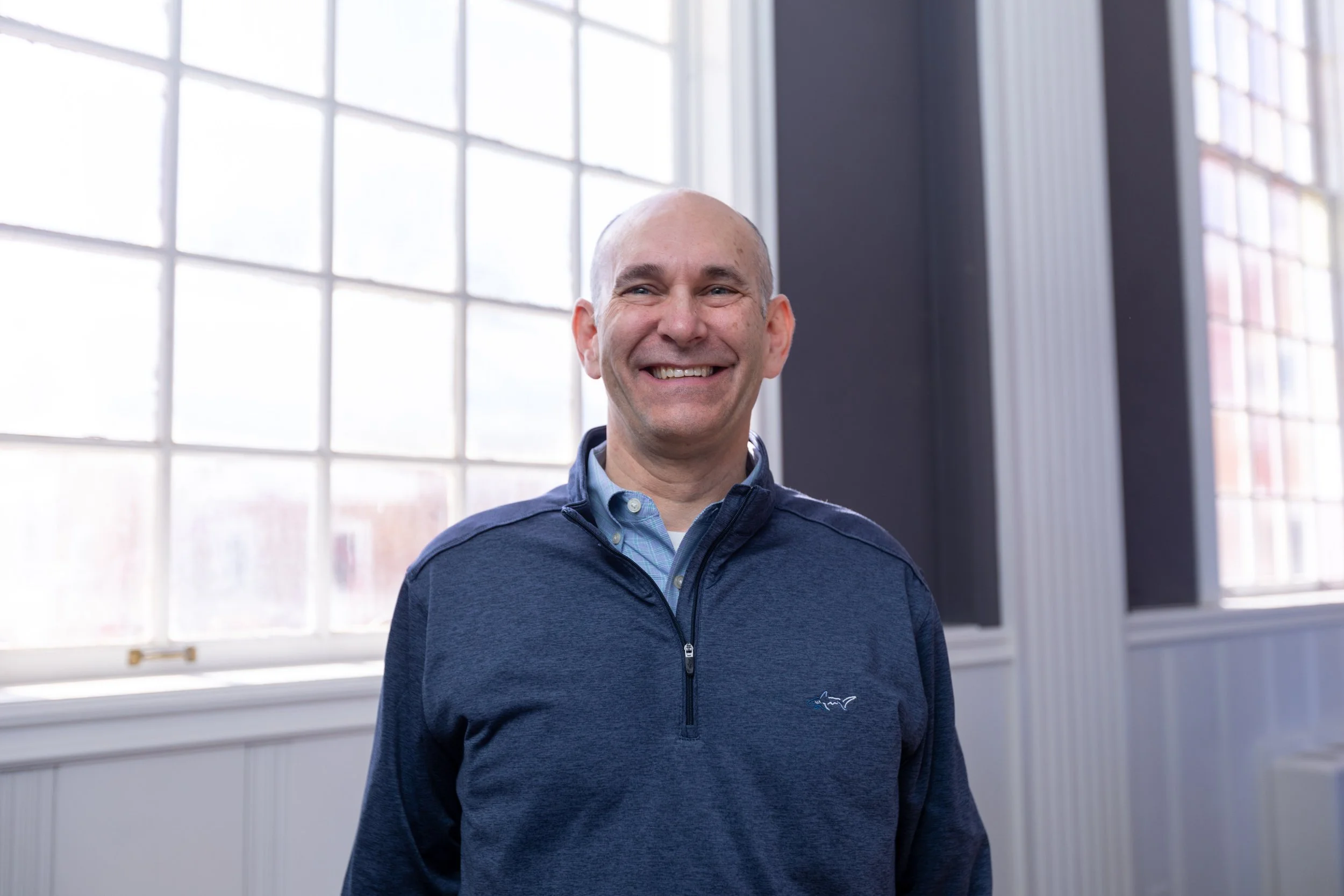 A middle-aged man with a shaved head wearing a blue Zip-up jacket smiling in front of large windows with natural light.