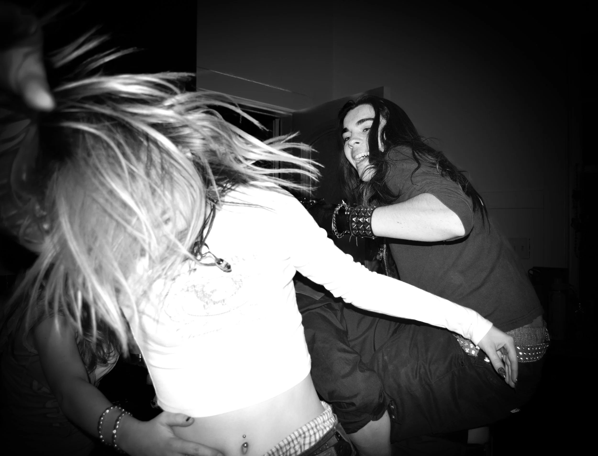 Two young women dancing energetically, one with dark hair smiling, the other with blond hair with her head down, captured in black and white