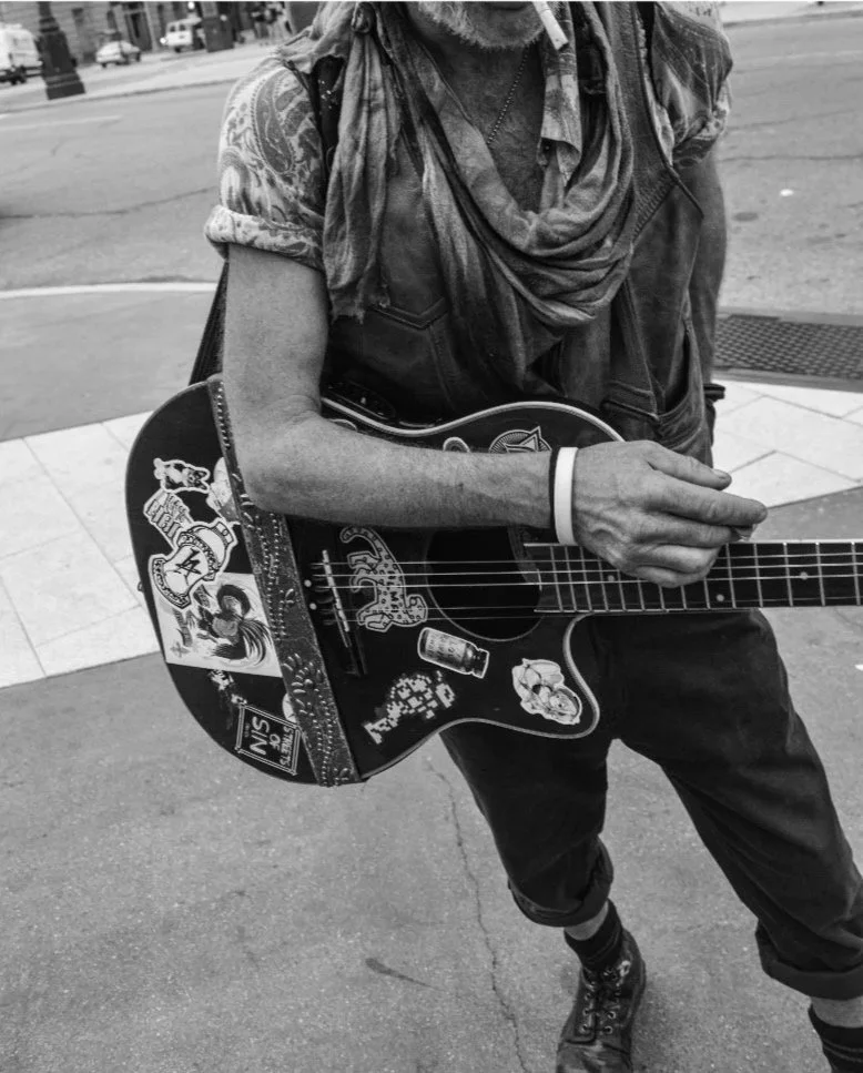 A person playing an acoustic guitar outdoors, decorated with various stickers, wearing a scarf and a vest, with tattoos visible on their arms.