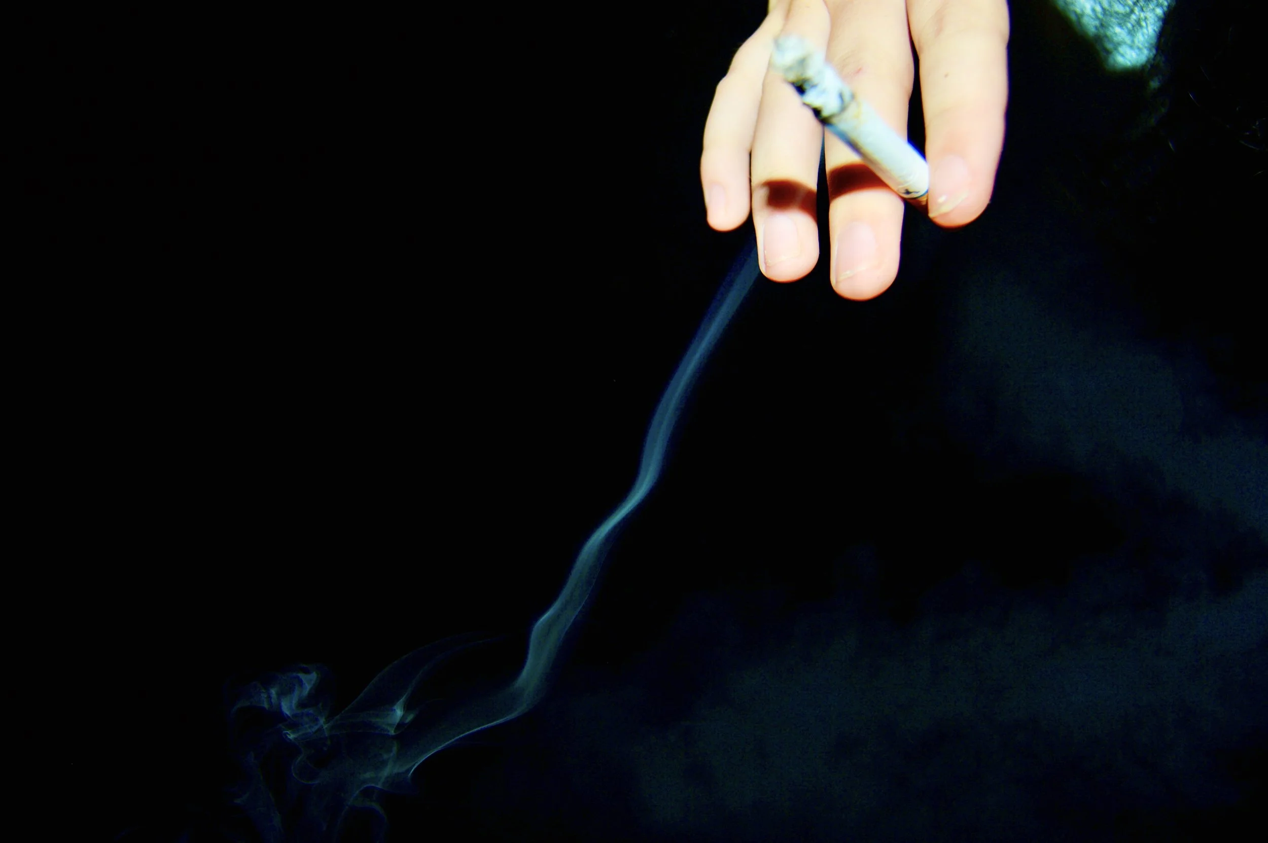 A hand holding a lit cigarette, with smoke rising against a dark background.