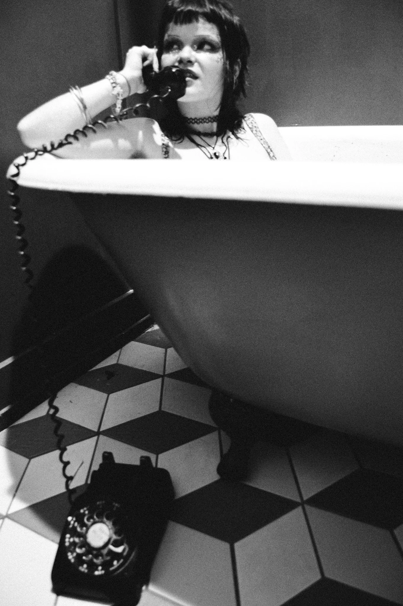 A person with dark hair and dark makeup sitting in a bathtub, talking on a corded phone, with a necklace and bracelets, in a room with patterned floor tiles.