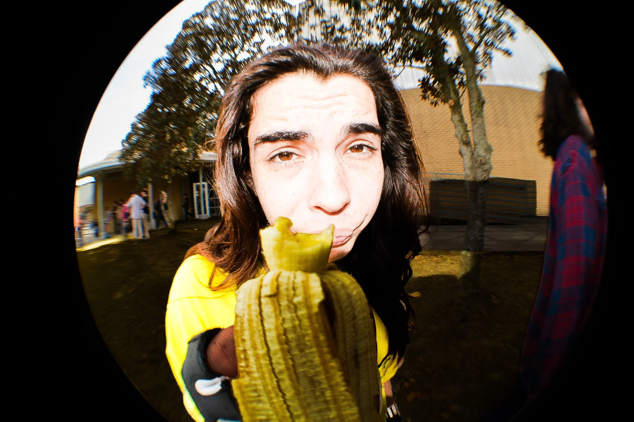 Close-up of a teenage girl with shoulder-length dark hair, wearing a yellow jacket and a yellow knitted glove, holding a pickle close to the camera lens. She is outdoors, with other people and a school building in the background, and trees are visible. The photo is taken with a fisheye lens, creating a circular, distorted perspective.