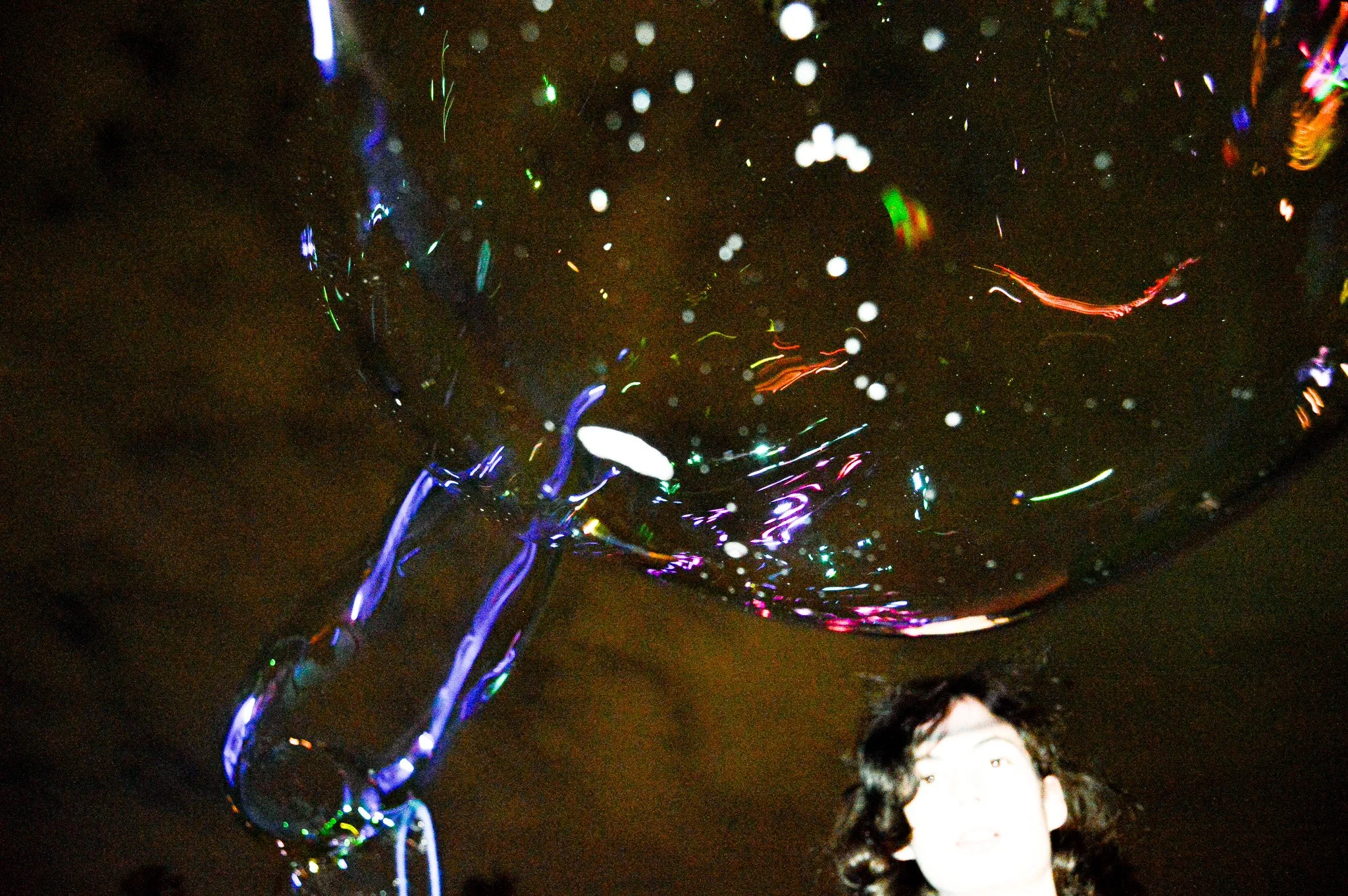 Person with dark curly hair standing beneath a large soap bubble with colorful reflections, against a dark background.