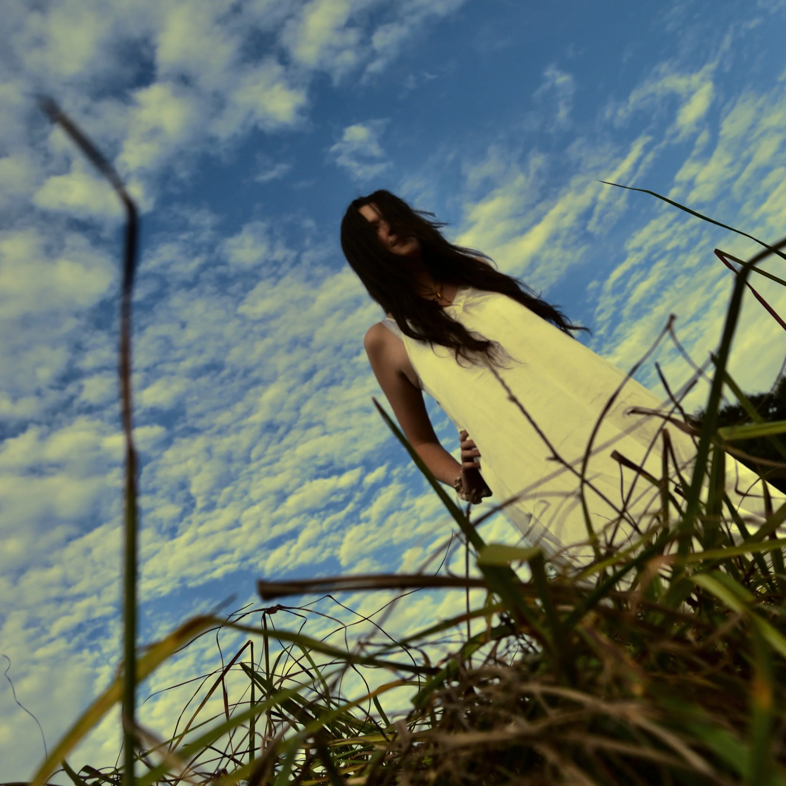A woman in a yellow dress stands outdoors among tall grass, with a bright blue sky filled with scattered clouds in the background.