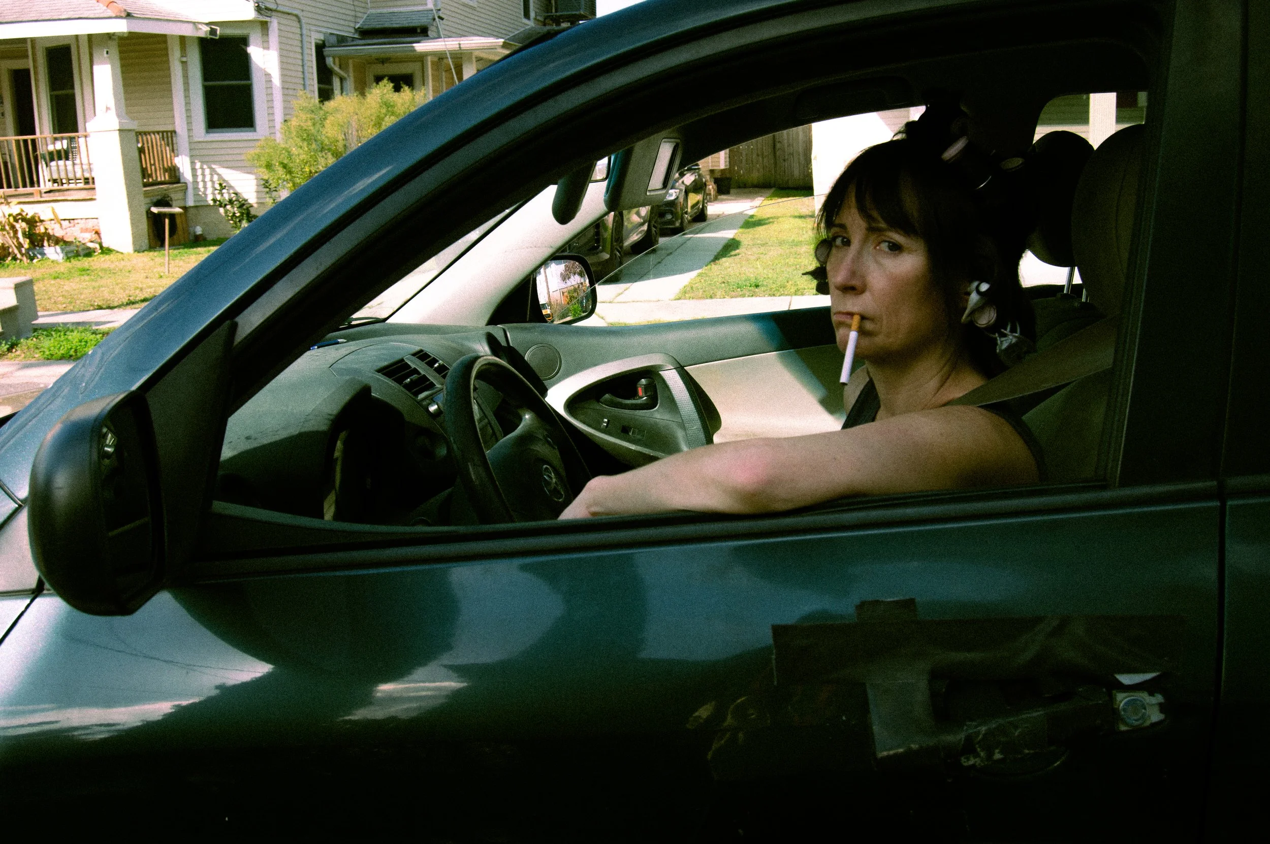 A woman with dark hair, wearing a black shirt and AirPods, sitting in the driver’s seat of a car, smoking a cigarette, with her arm resting on the window frame, in a residential neighborhood.