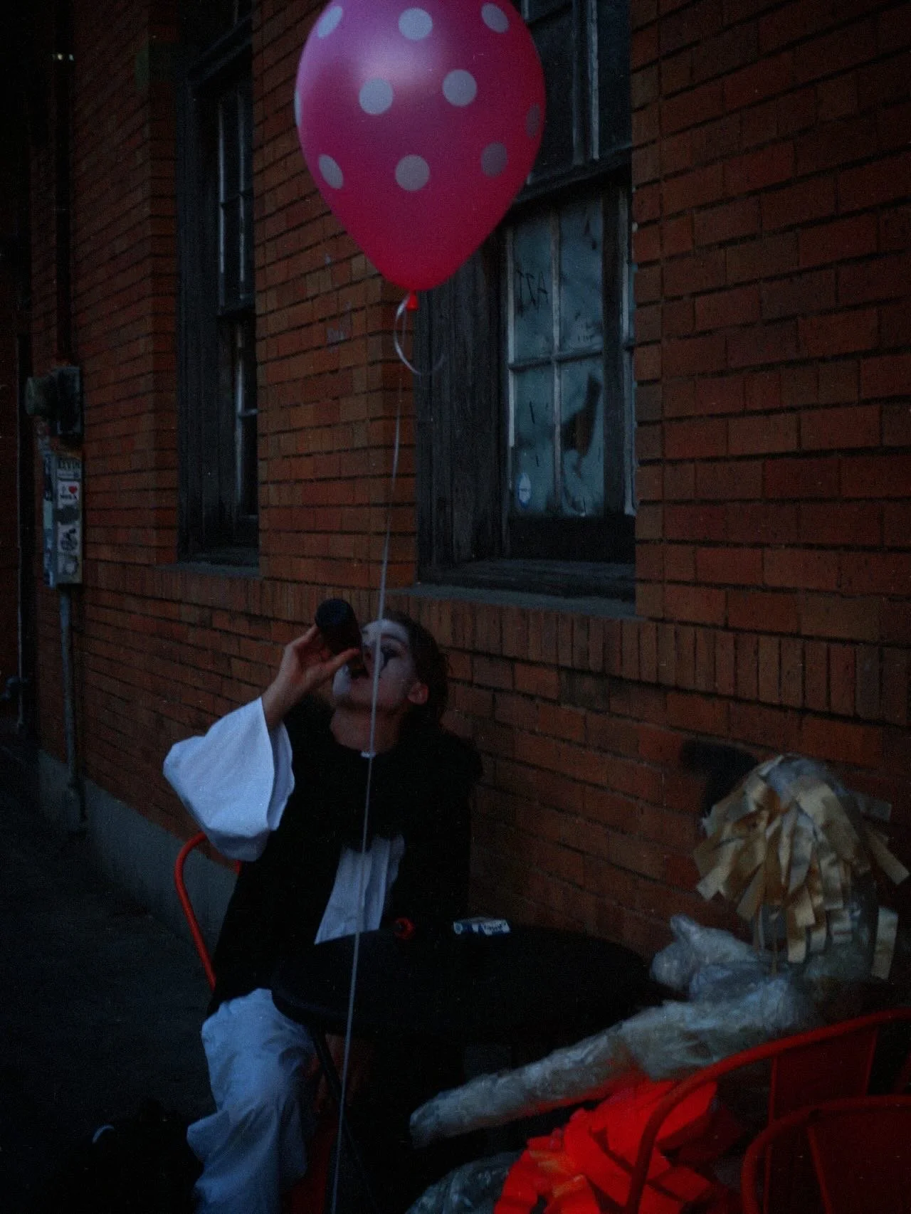 Person dressed in clown makeup and costume sitting outdoors next to a brick building, holding a pink polka-dot balloon, drinking from a bottle.