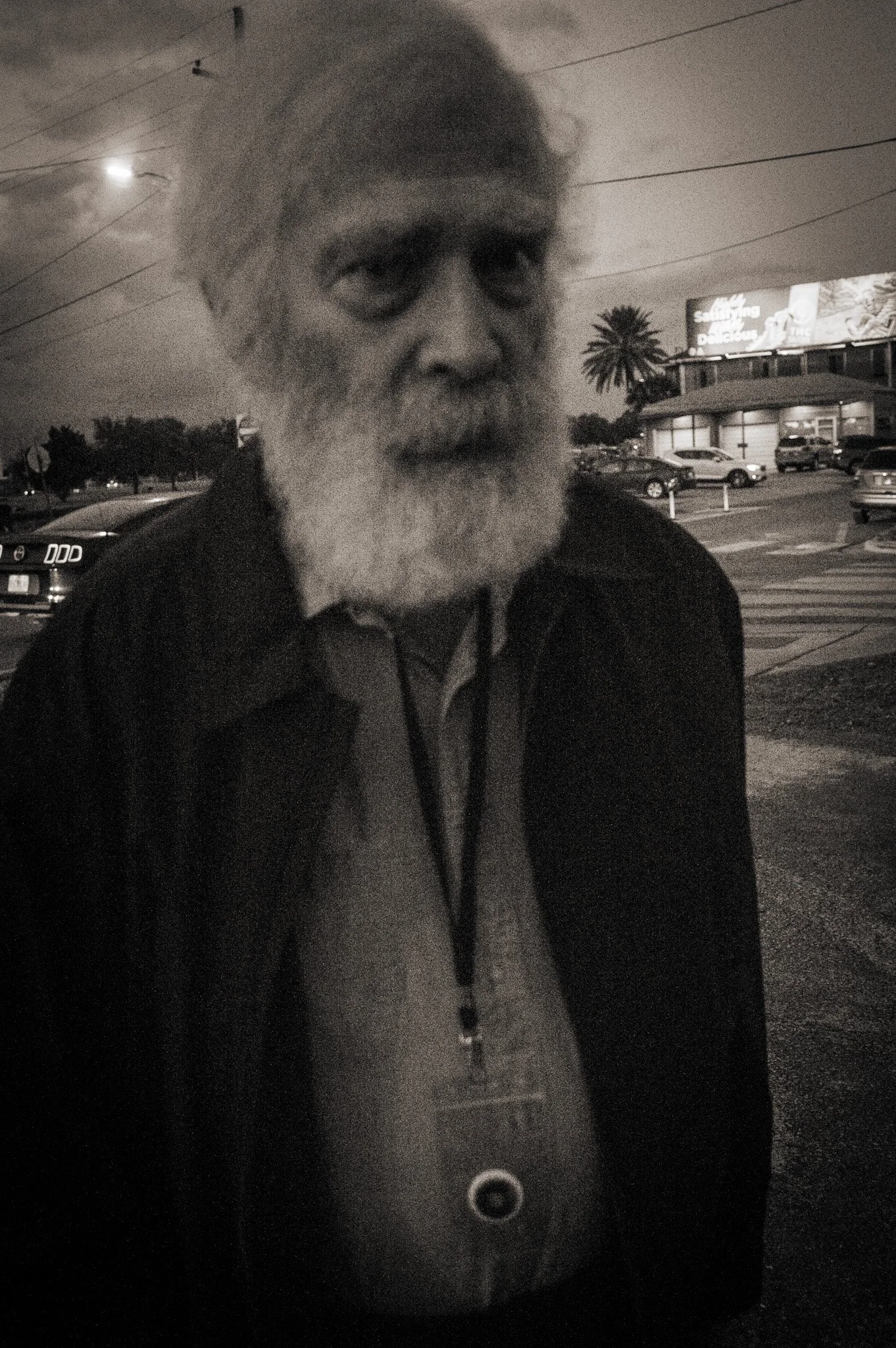 An older man with a white beard and hair, wearing a dark jacket and a lanyard with a badge, standing outdoors in a parking lot at night.