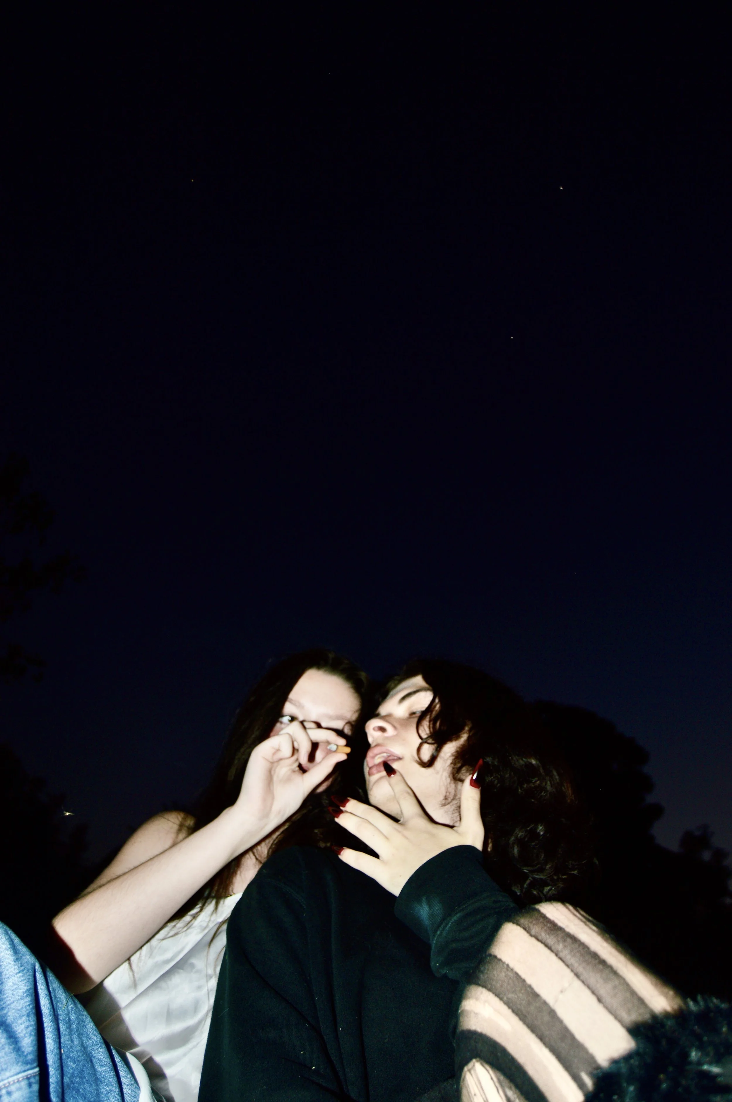 Two women at night, one with glasses and the other with dark curly hair, posing closely with one woman touching her face and the other holding a cigarette.
