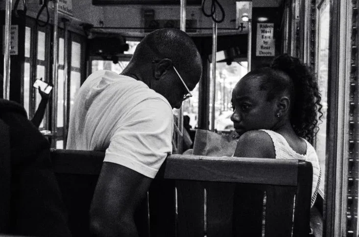 A man and woman sitting together on a bus, engaged in conversation, with the man wearing glasses and a white T-shirt, and the woman with curly hair tied back.