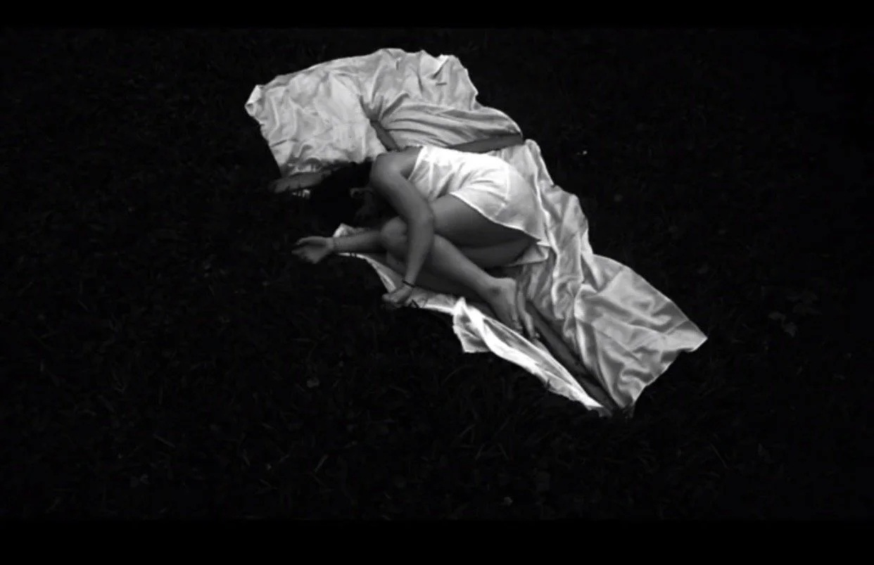 A person sleeping curled up on a dark outdoor surface, covered with a white blanket and wearing a white sleeveless top and shorts.