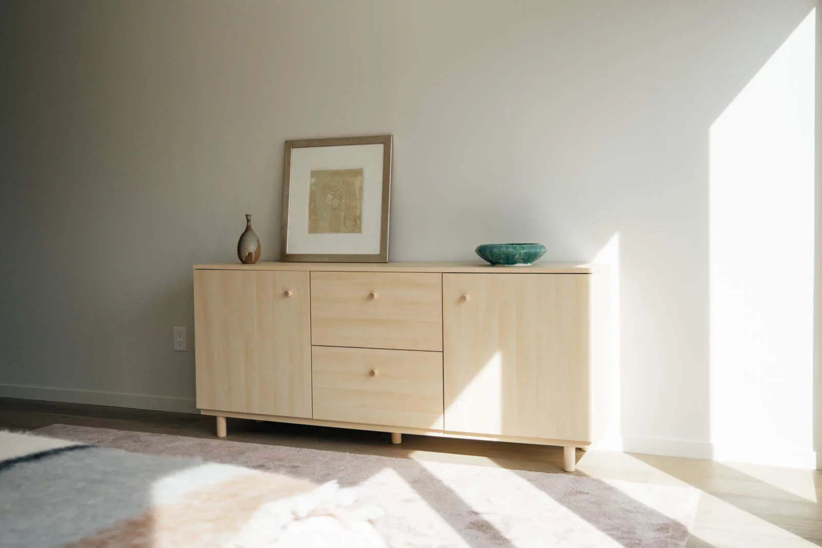 Light-colored wooden sideboard with three drawers and two cabinets, decorated with a small vase, a framed picture, and a green bowl, in a sunlit room with beige walls and a multicolored rug.