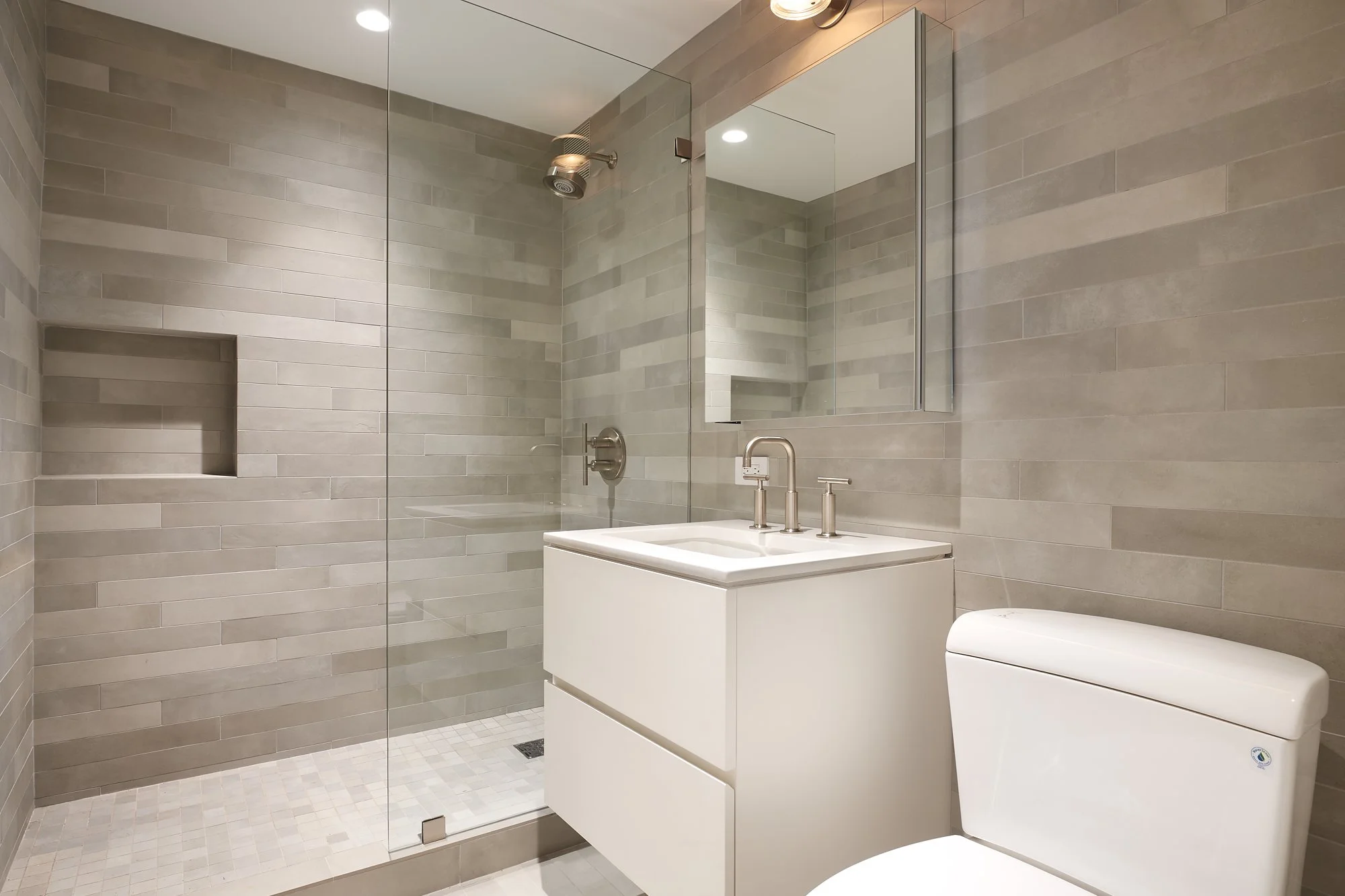 Modern bathroom with walk-in shower, neutral-toned tiles, a white vanity with a sink and faucet, a wall-mounted mirror, and a toilet.