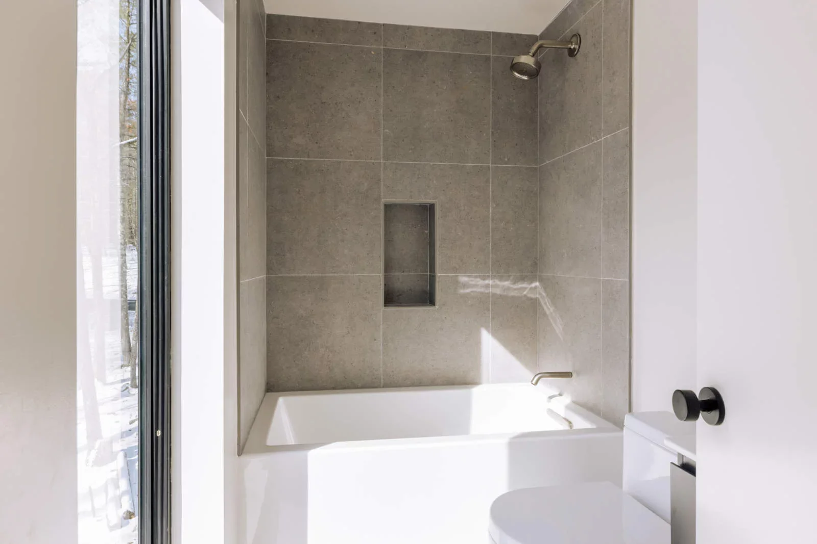 A modern bathroom with a bathtub, gray tiled wall, built-in shower niche, and a ceiling shower head. A glass door and a window are partially visible, allowing natural light.